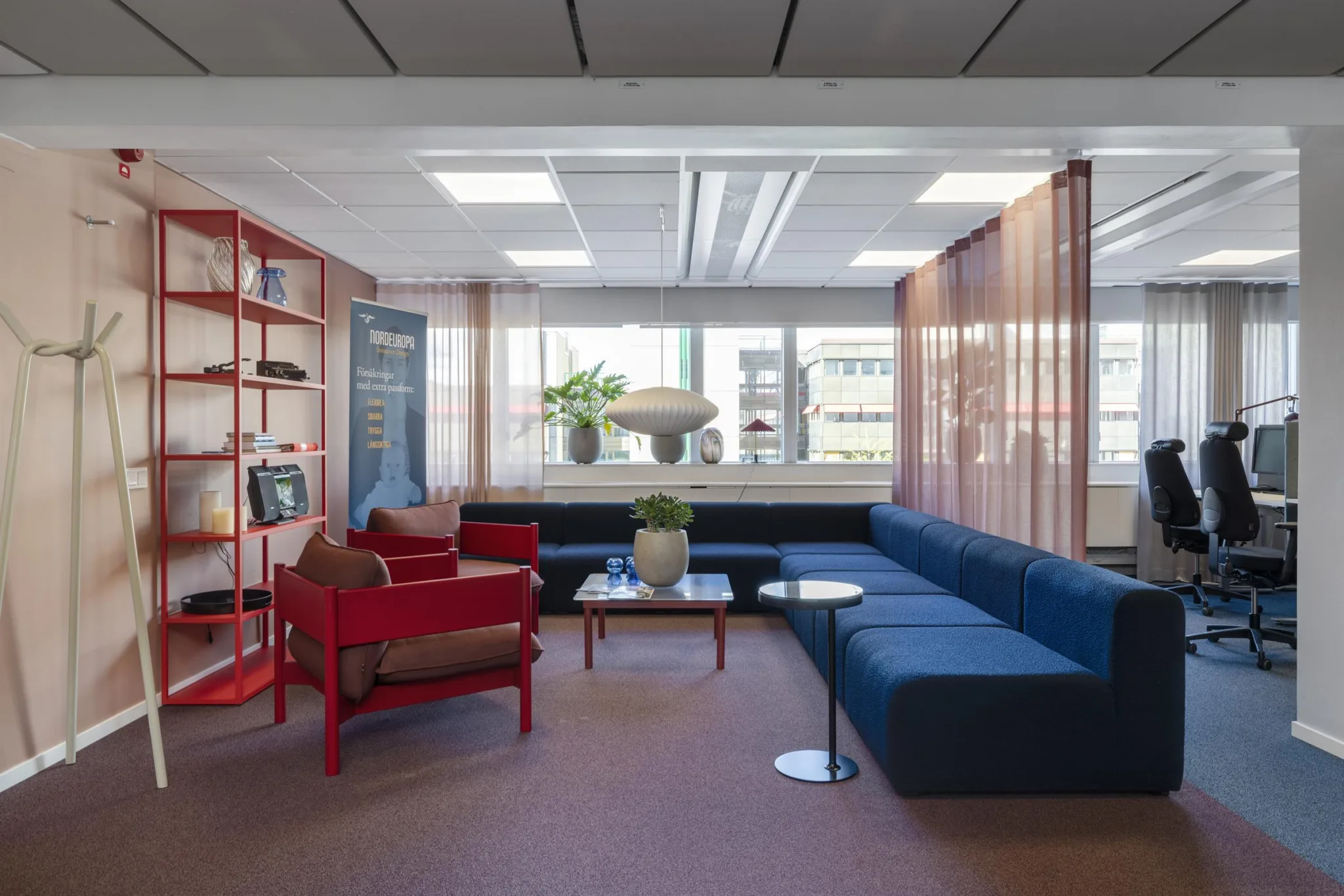5. Ett loungeområde med röda och blå soffor, böcker på hyllor, en stor krukväxt och fönster med gardiner.