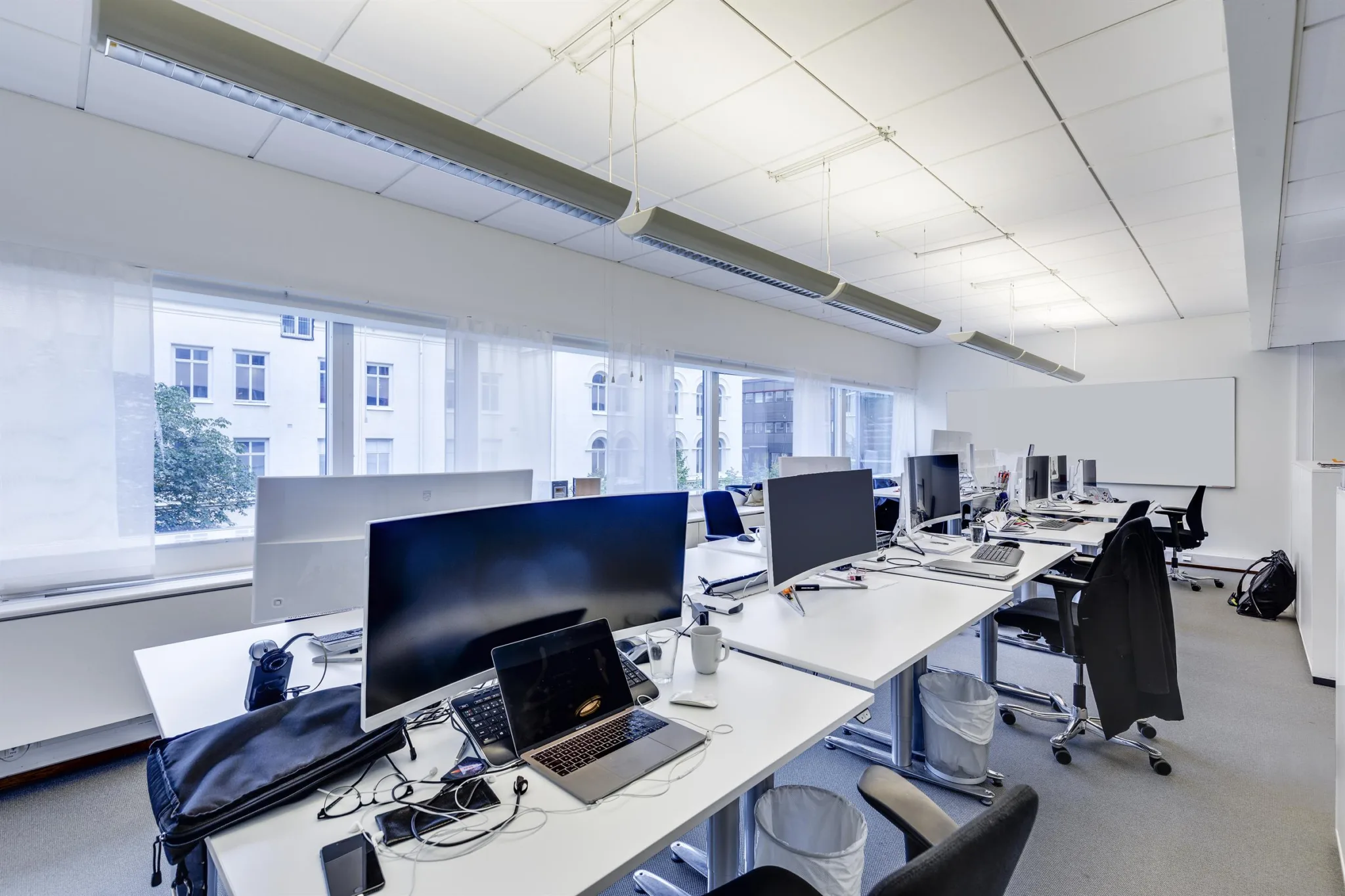 En öppen kontorsyta med långa skrivbord fyllda med datorskärmar, bärbara datorer och tillbehör, samt stora fönster i bakgrunden.