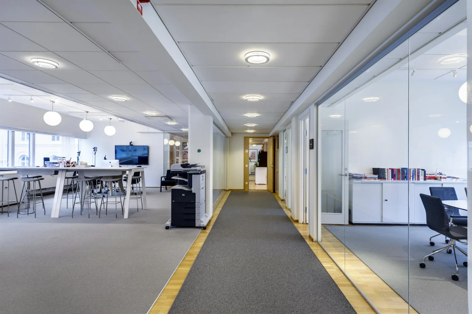 En lång korridor med grå matta, kontorsrum på höger sida och ett öppet mötesområde till vänster.