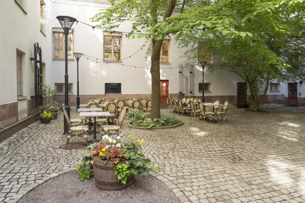 Innergård med kullersten, träd och utemöbler, omgiven av vita byggnader och dekorerad med blommor.