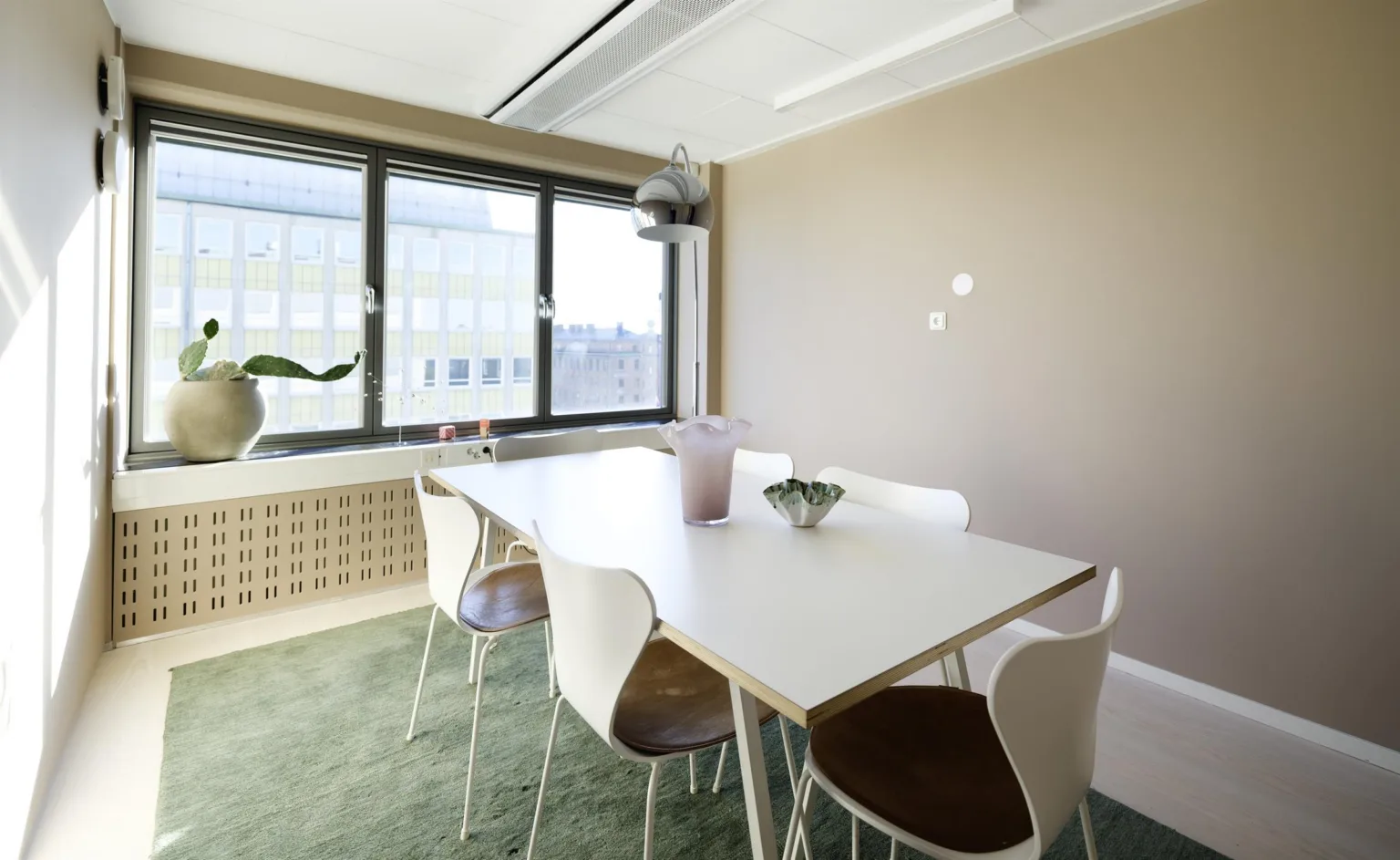Mötesrum med ett vitt bord, fyra stolar, växtdekorationer och stora fönster med utsikt.