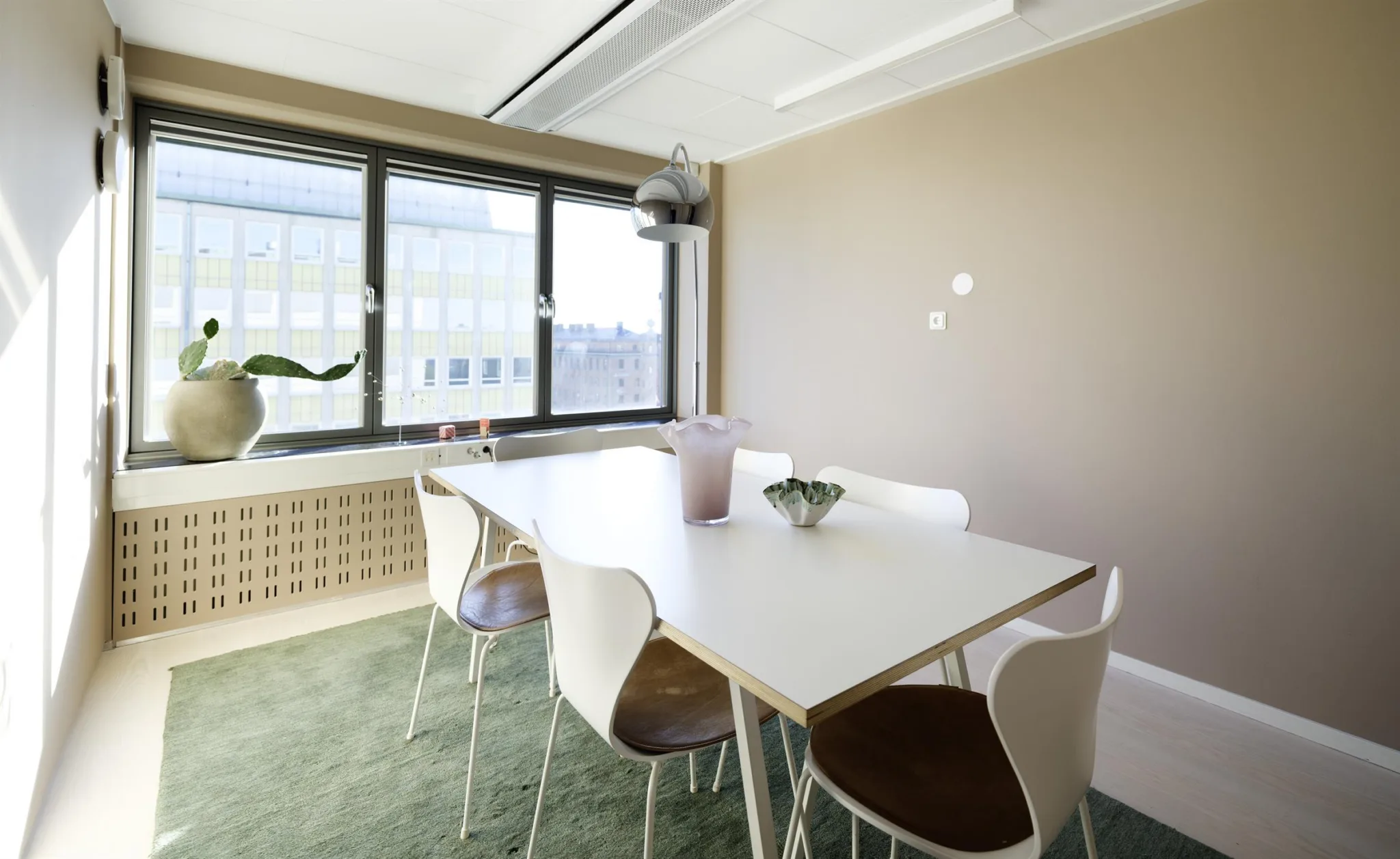 Mötesrum med ett vitt bord, fyra stolar, växtdekorationer och stora fönster med utsikt.