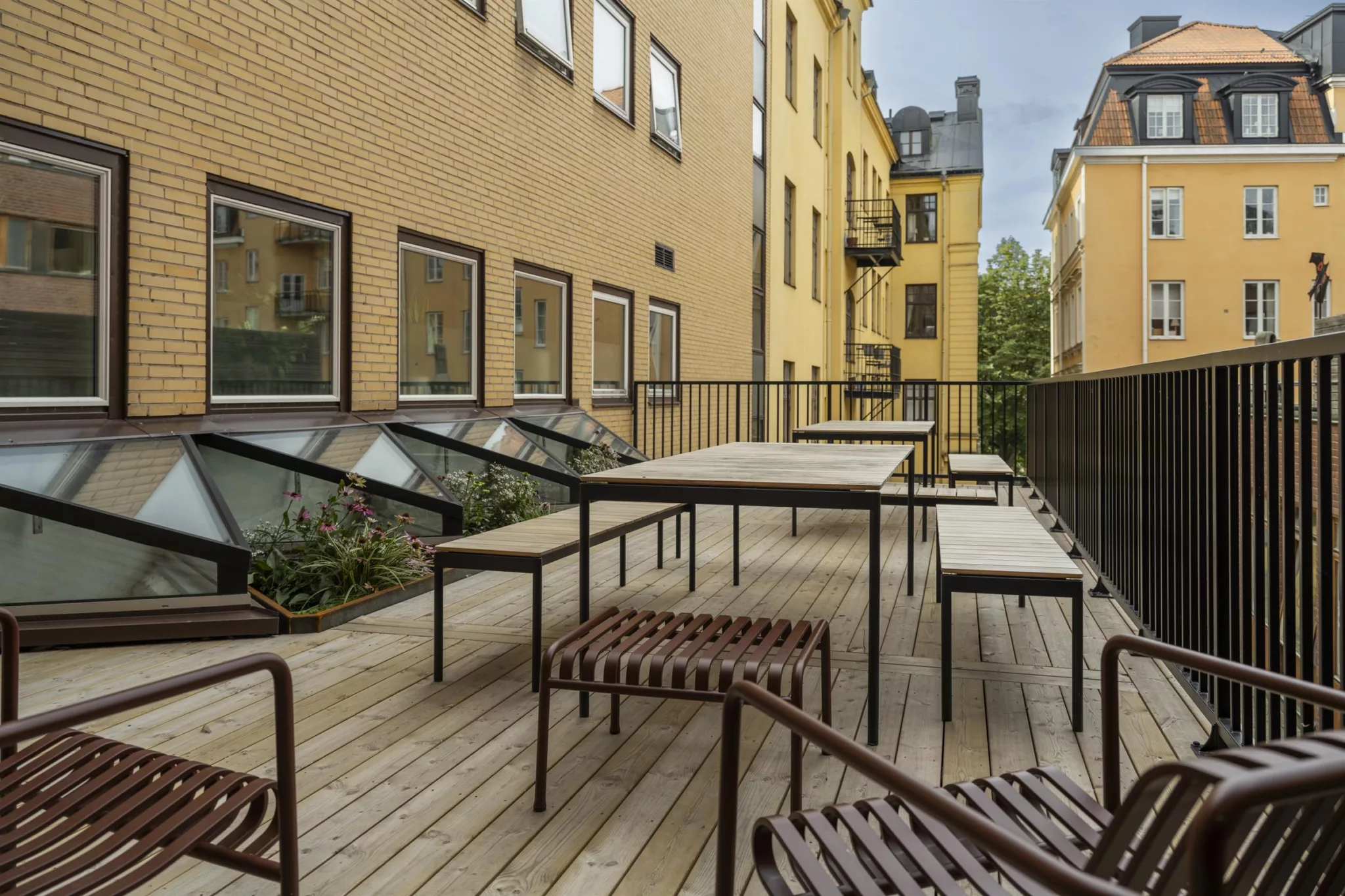 En träterrass med bord och stolar omgiven av gula fasader och fönster.