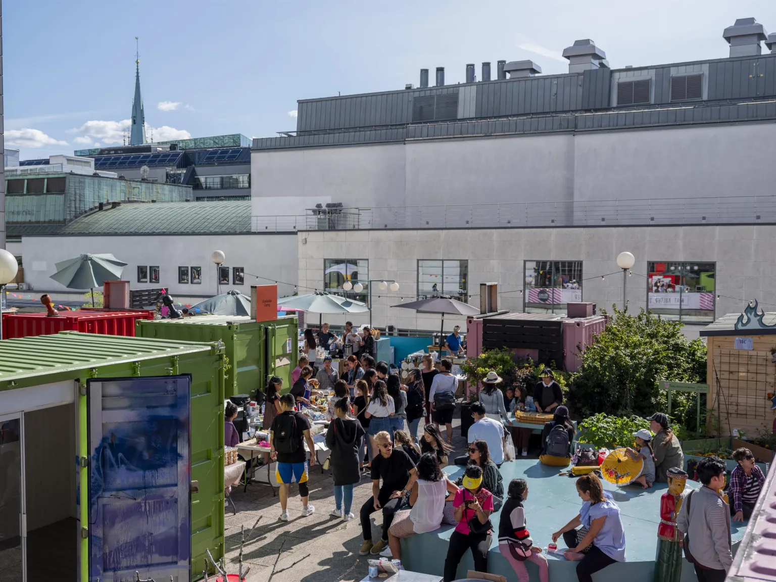 2. En livlig utomhusyta med människor på bänkar och sittplatser, omgiven av färgglada containrar och en basketplan.