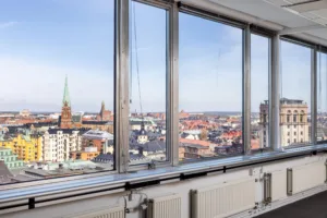 2. Utsikt över en stad med kyrktorn och byggnader, sett genom stora glasfönster från ett högt beläget rum.