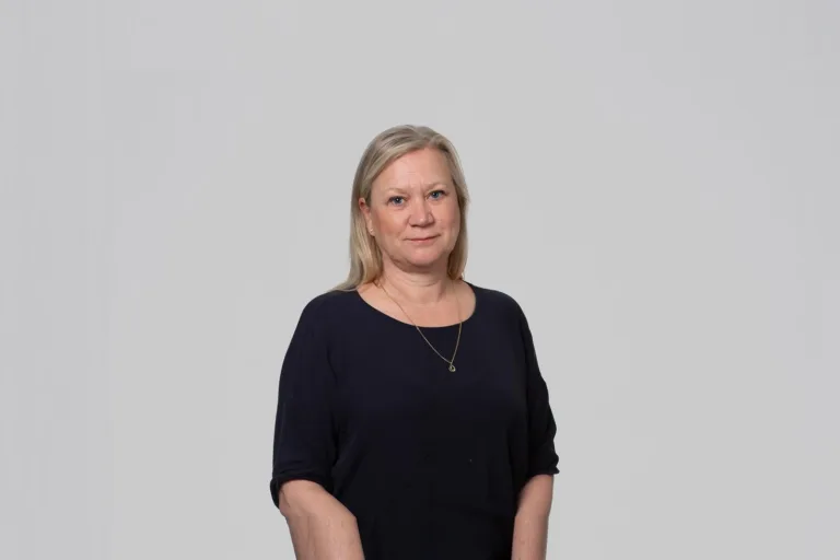 En kvinna med kort blont hår, klädd i en svart topp, står med avslappnad hållning mot en ljus bakgrund.