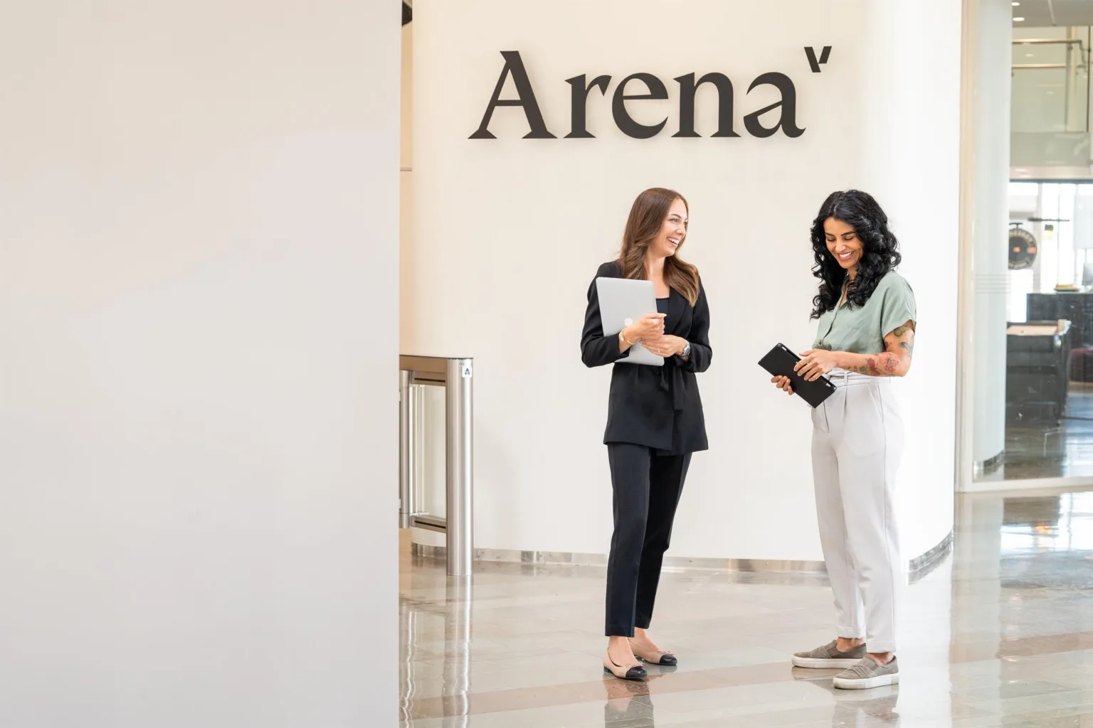 Två kvinnor står framför en vägg med texten "Arena"; en håller dokument och den andra håller en telefon.