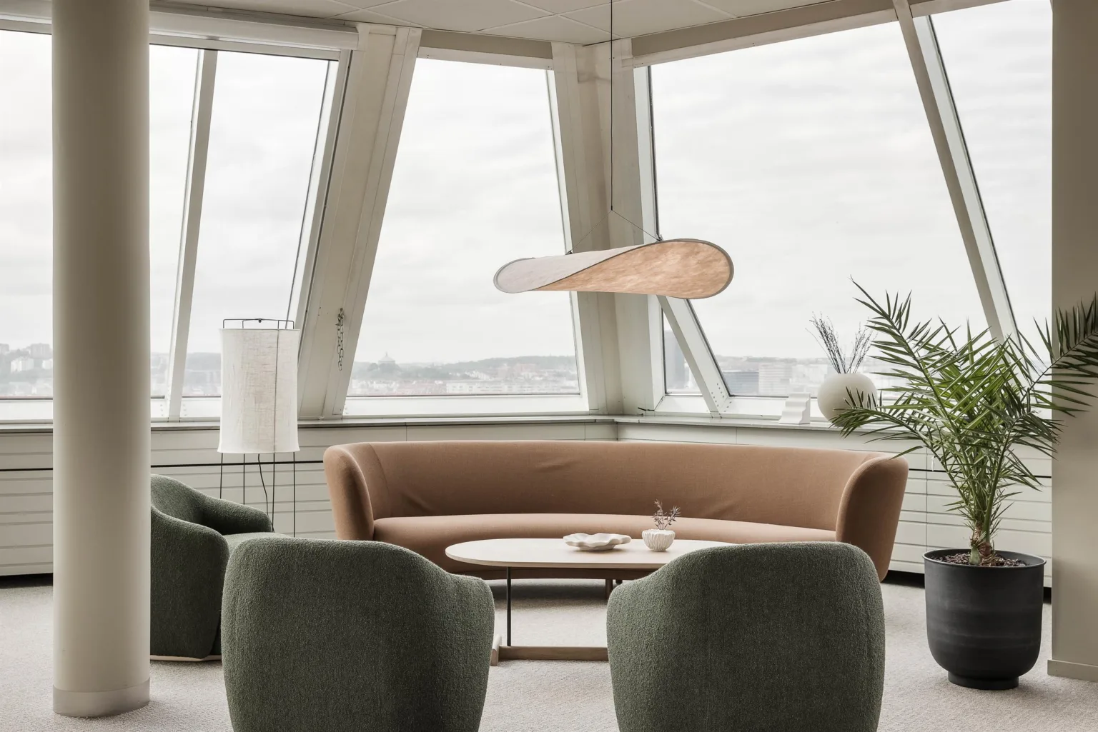 En stilren lounge med en beige soffa, gröna fåtöljer och ett modernt hängande ljus framför stora fönster.