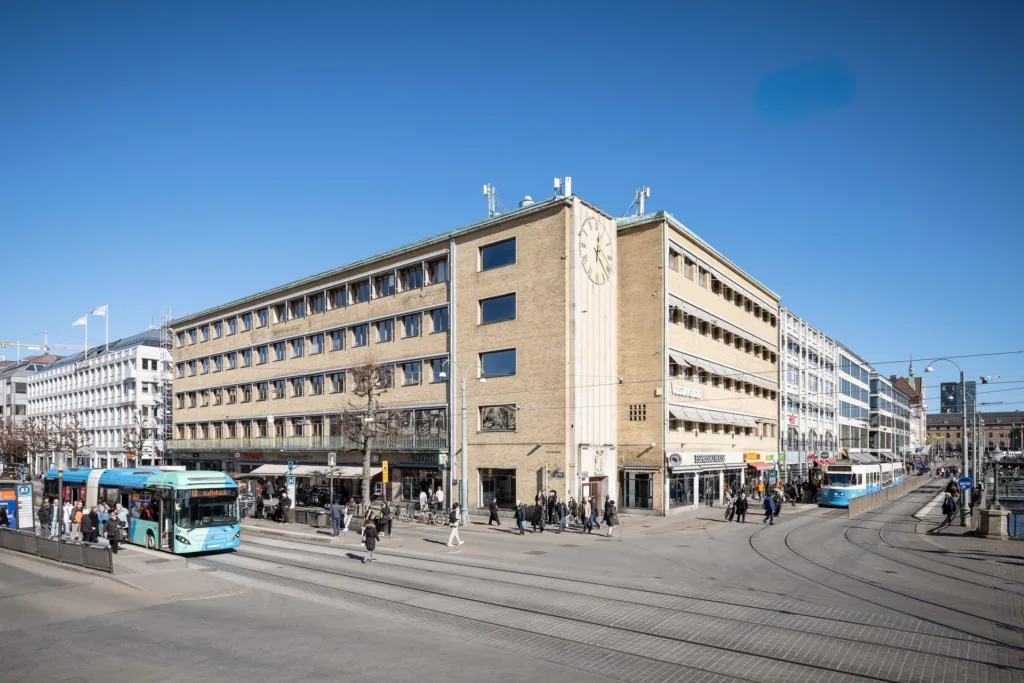 En stor byggnad i tegel vid en trafikerad gata, med människor, spårvagnar och cyklister i rörelse.