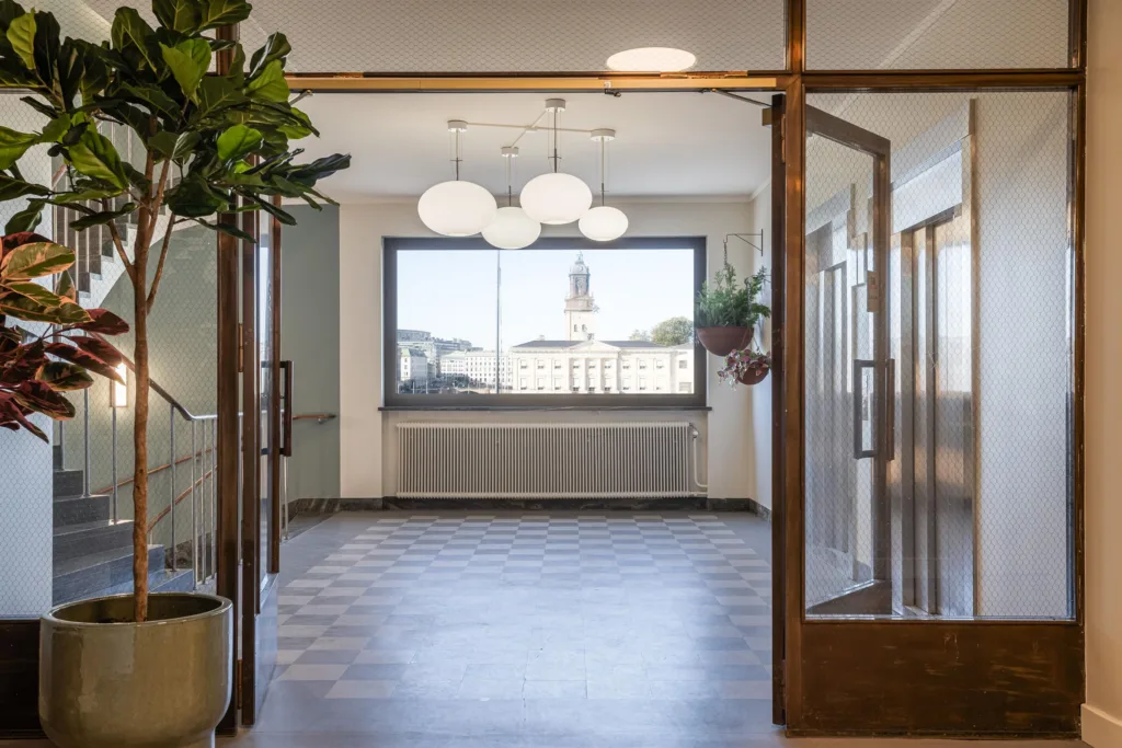 En korridor med glasdörrar, stora fönster och hängande taklampor, med utsikt över ett kyrktorn i fjärran.