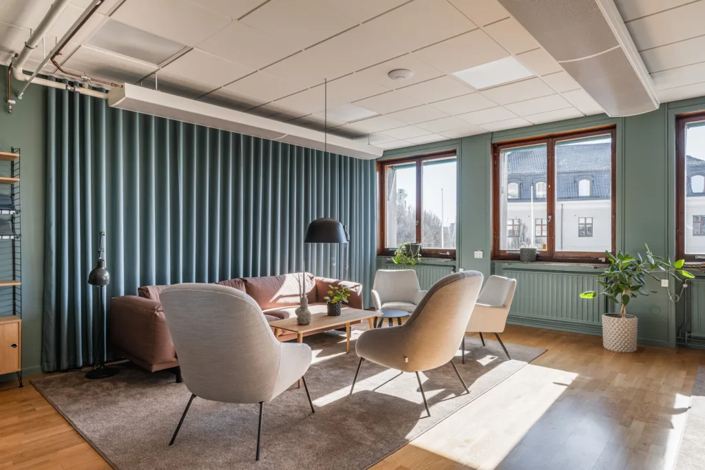 1. Ett modernt kontorsutrymme med fåtöljer, soffa och bord, omgiven av gröna väggar och stora fönster som släpper in dagsljus.