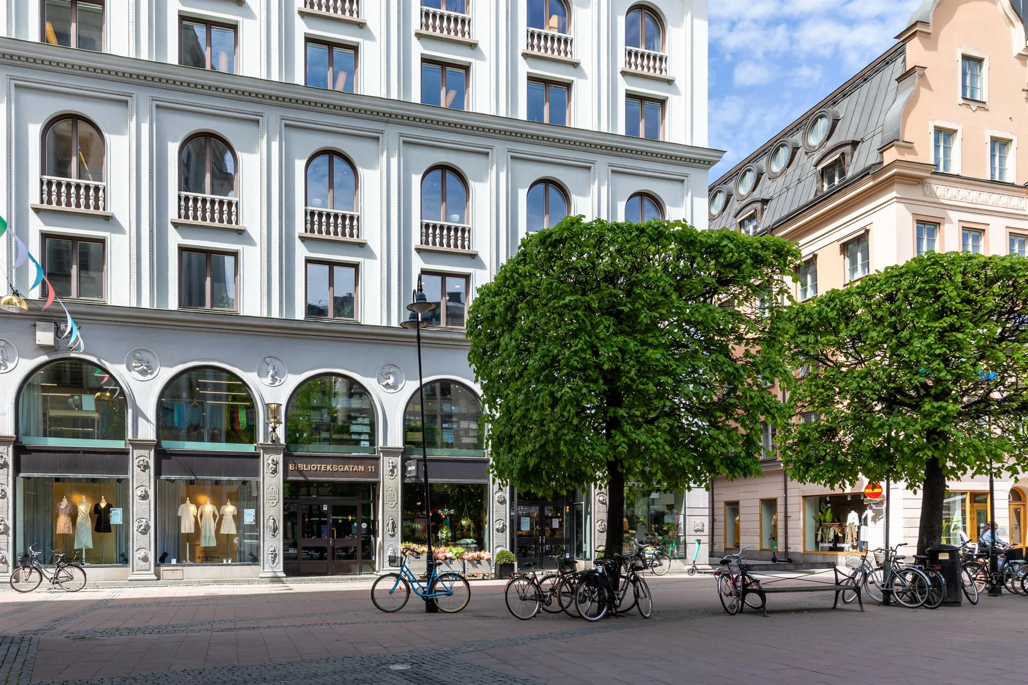 En vit byggnad med bågformade fönster, cyklar parkerade framför och ett stort träd vid sidan.