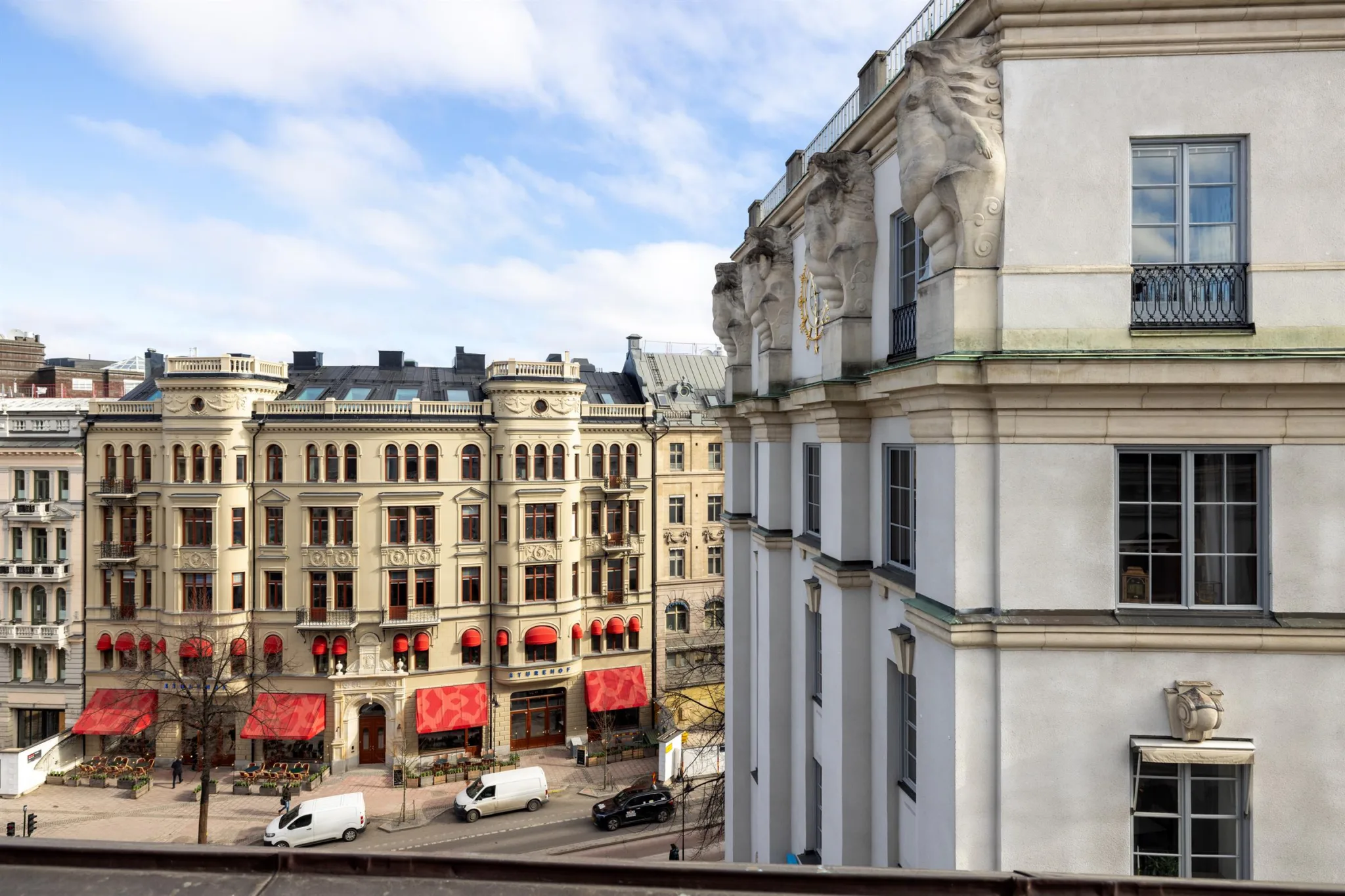 Stadsvy med klassiska byggnader, röda markiser och bilar på gatan under en klar himmel.