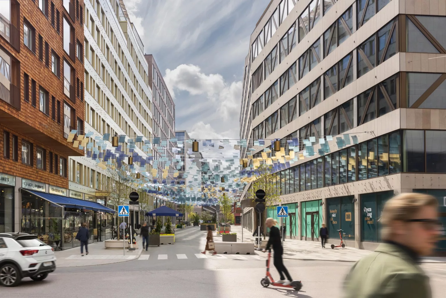 En stadsgata med moderna byggnader på båda sidor, dekorerad med färgglada hängande band, och en person som åker elsparkcykel.