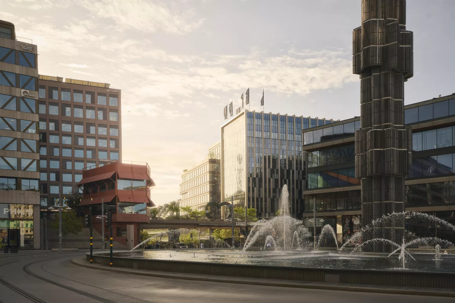 En fontän omgiven av byggnader i olika stilar, med en klar himmel och solnedgång som skapar en varm atmosfär.