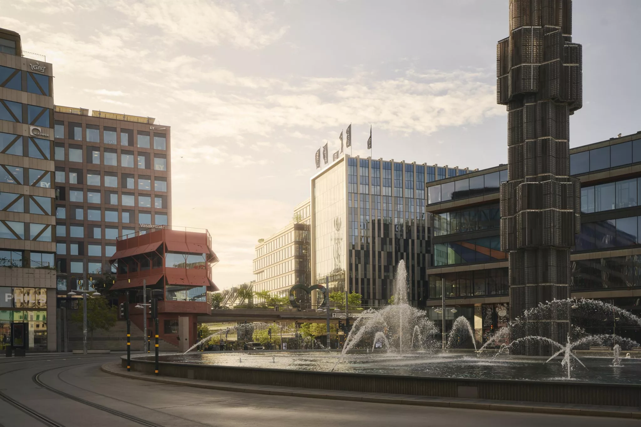 En fontän omgiven av byggnader i olika stilar, med en klar himmel och solnedgång som skapar en varm atmosfär.