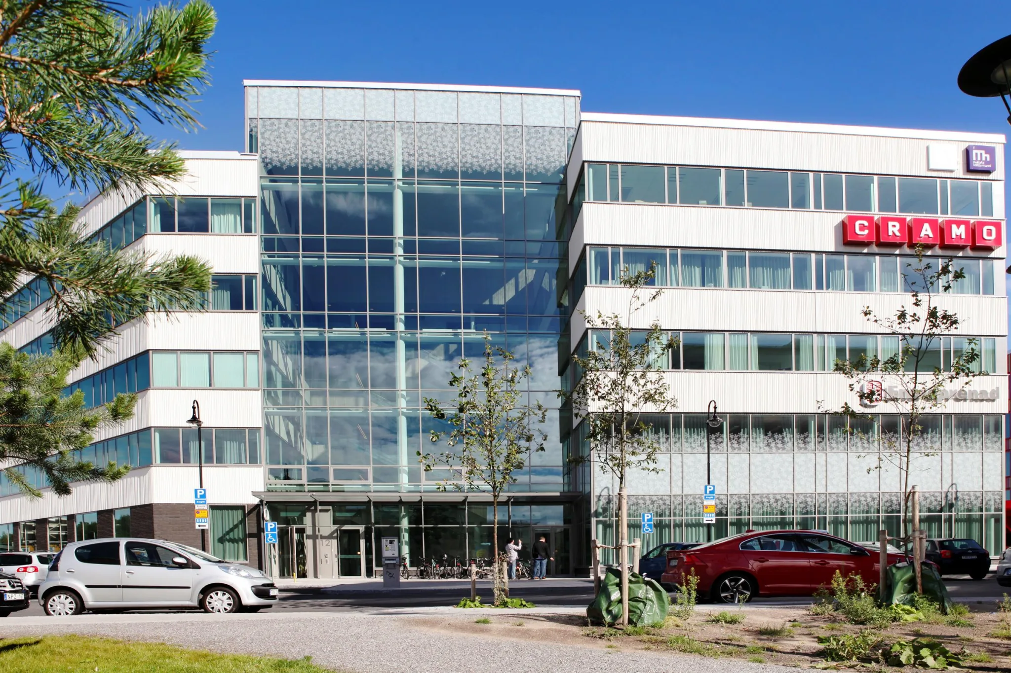En vit byggnad med glasfasad och parkerade bilar framför, omgiven av träd.