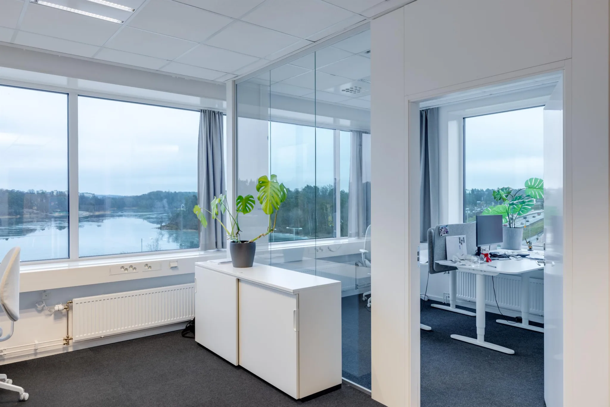 Ett kontor med glasväggar, vita möbler och utsikt över vatten och träd.