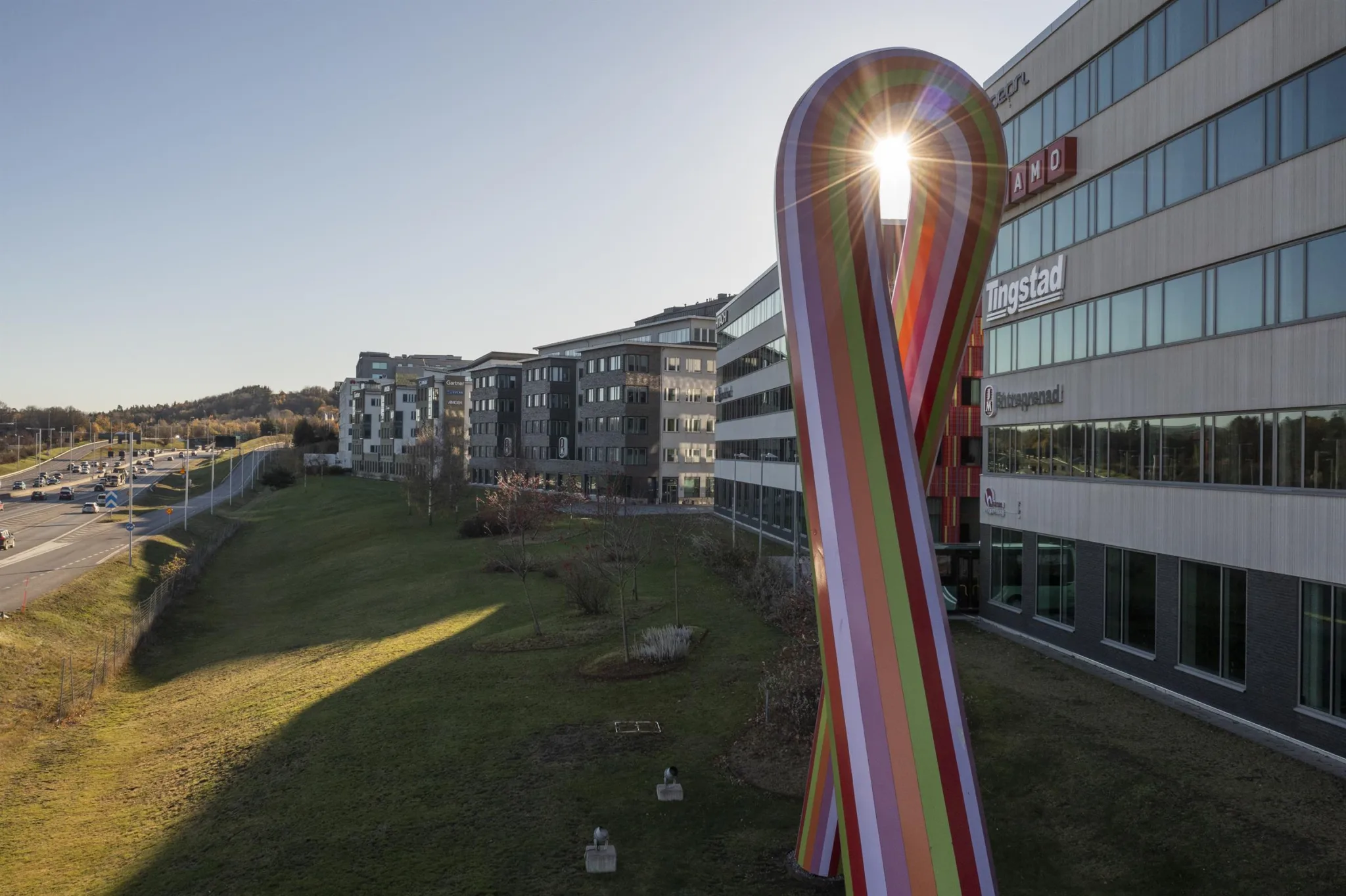 En stor, färgglad skulptur med solstrålar som lyser igenom, omgiven av byggnader och en grön gräsyta.
