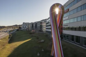 En stor, färgglad skulptur med solstrålar som lyser igenom, omgiven av byggnader och en grön gräsyta.