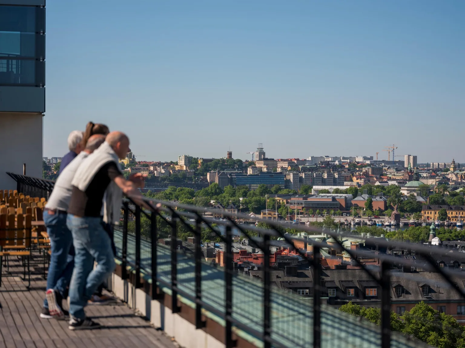 Tre personer står vid ett räcke och blickar ut över en stad med byggnader och grönska under en klar blå himmel.