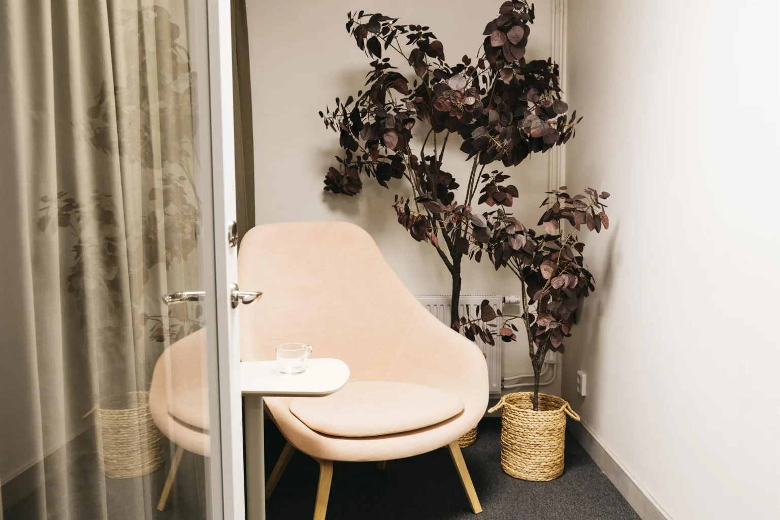 En liten avskild hörna med en ljusrosa stol, ett litet bord, och en dekorativ växt i en korg.