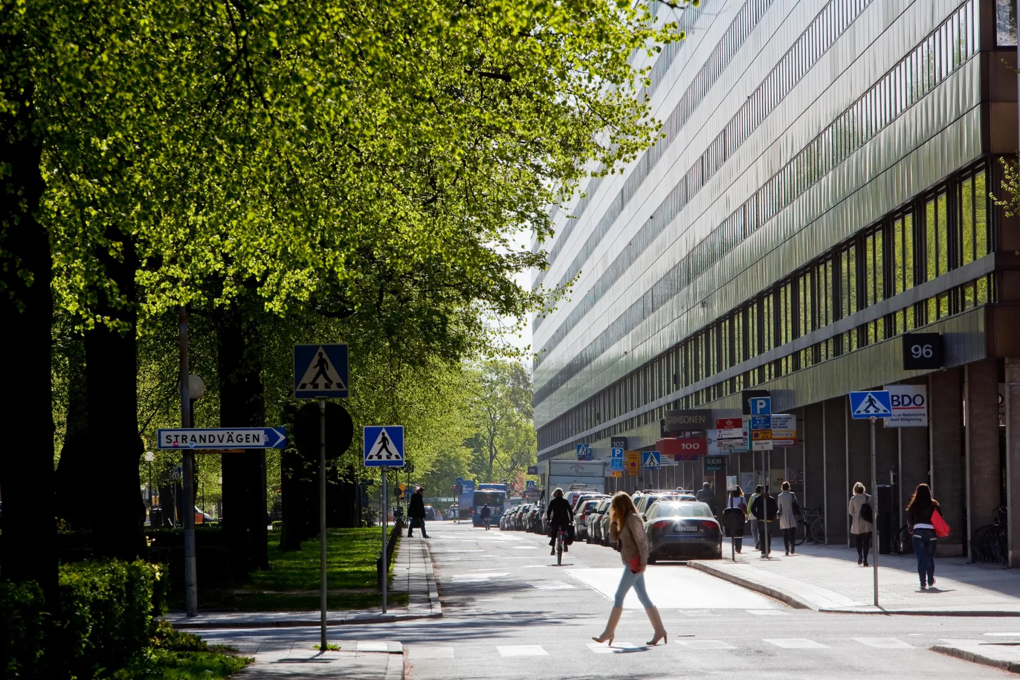 En stadsgata med grönska, en gångtrafikant och en lång byggnad med glasfasad.