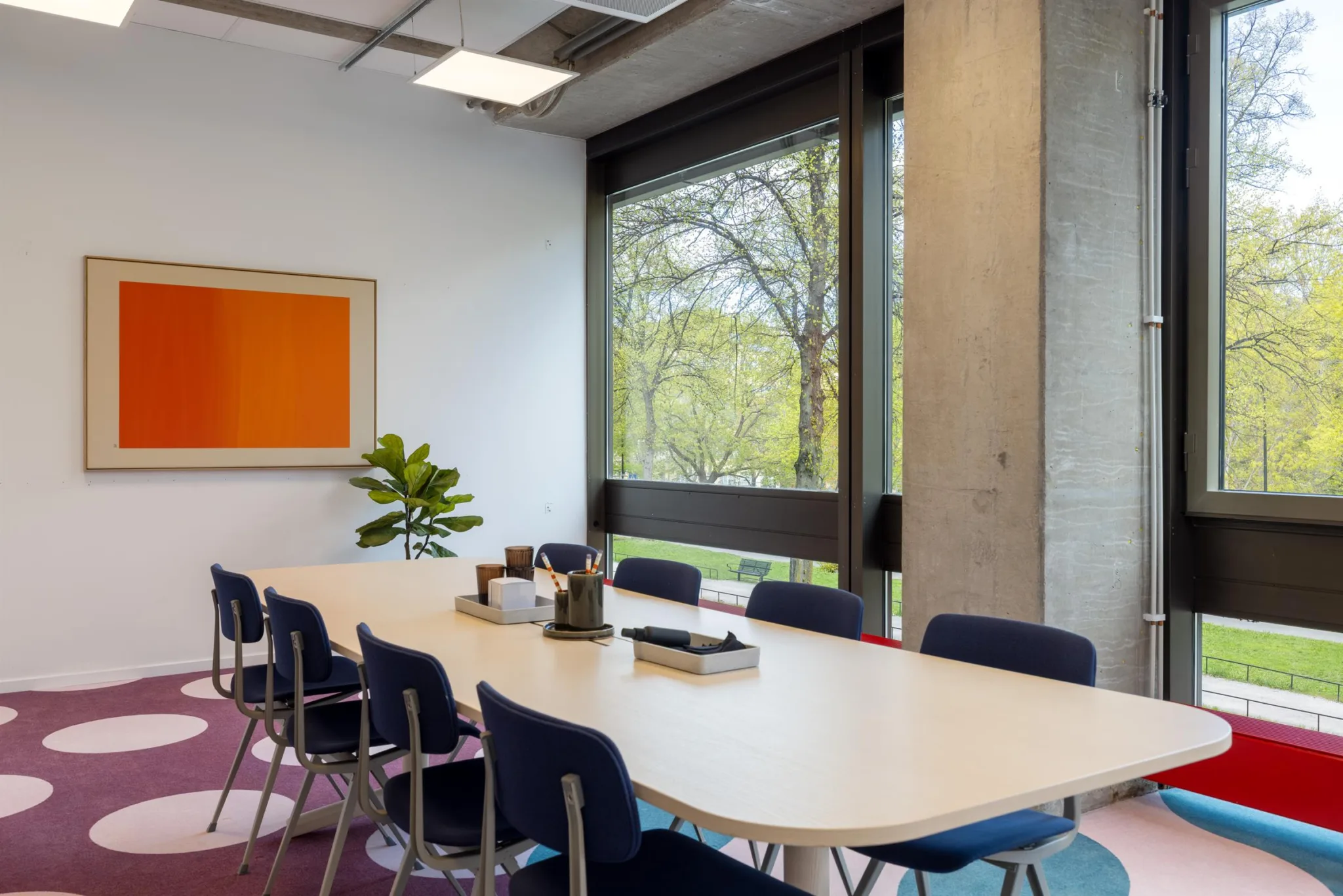 3. Konferensrum med lång bord, blåa stolar, växt och orange konstverk på väggen, stora fönster med utsikt över träd.