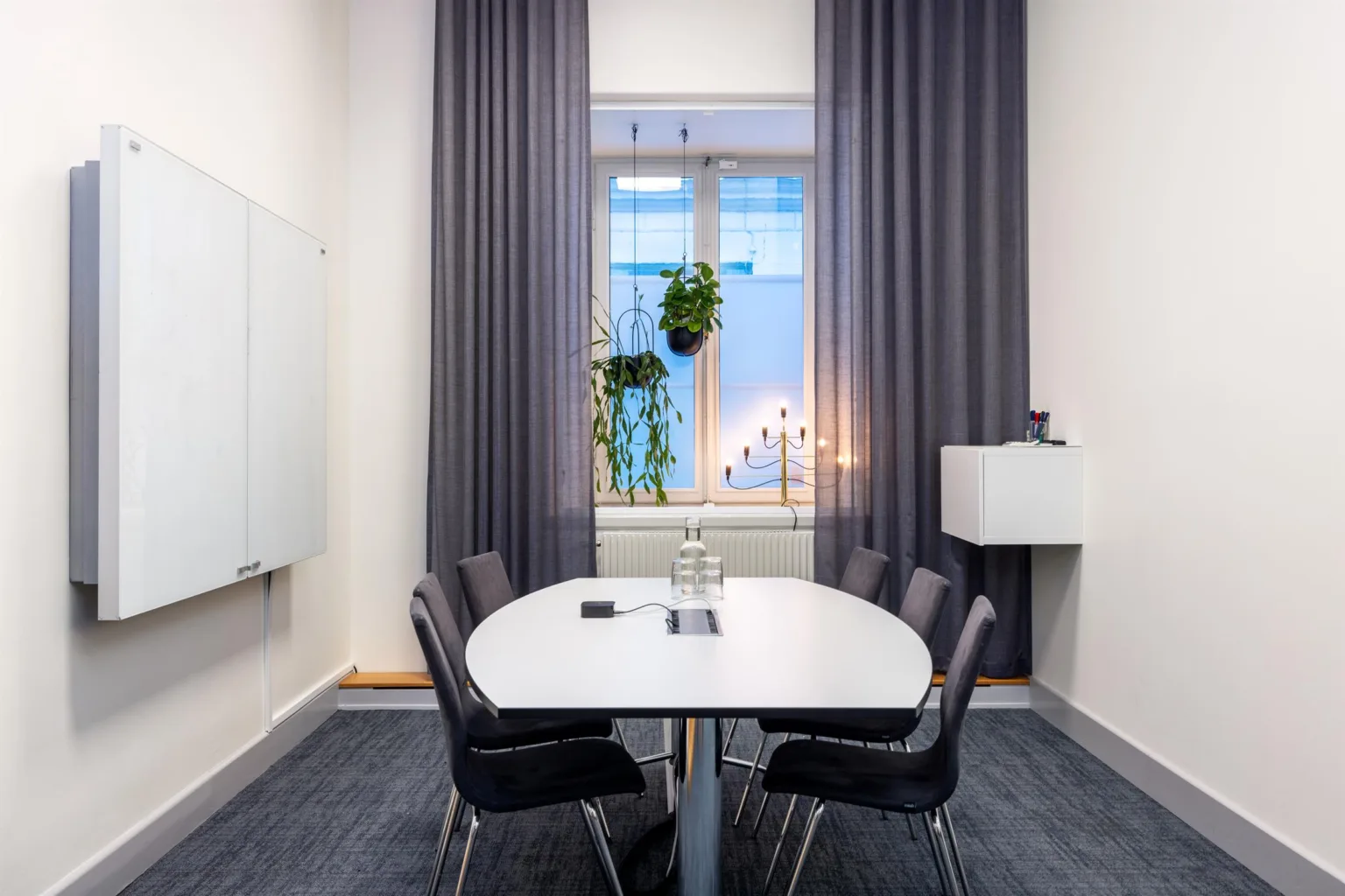 Ett mötesrum med ett vitt bord, fyra svarta stolar, mörka gardiner, ett fönster med växt och levande ljus.