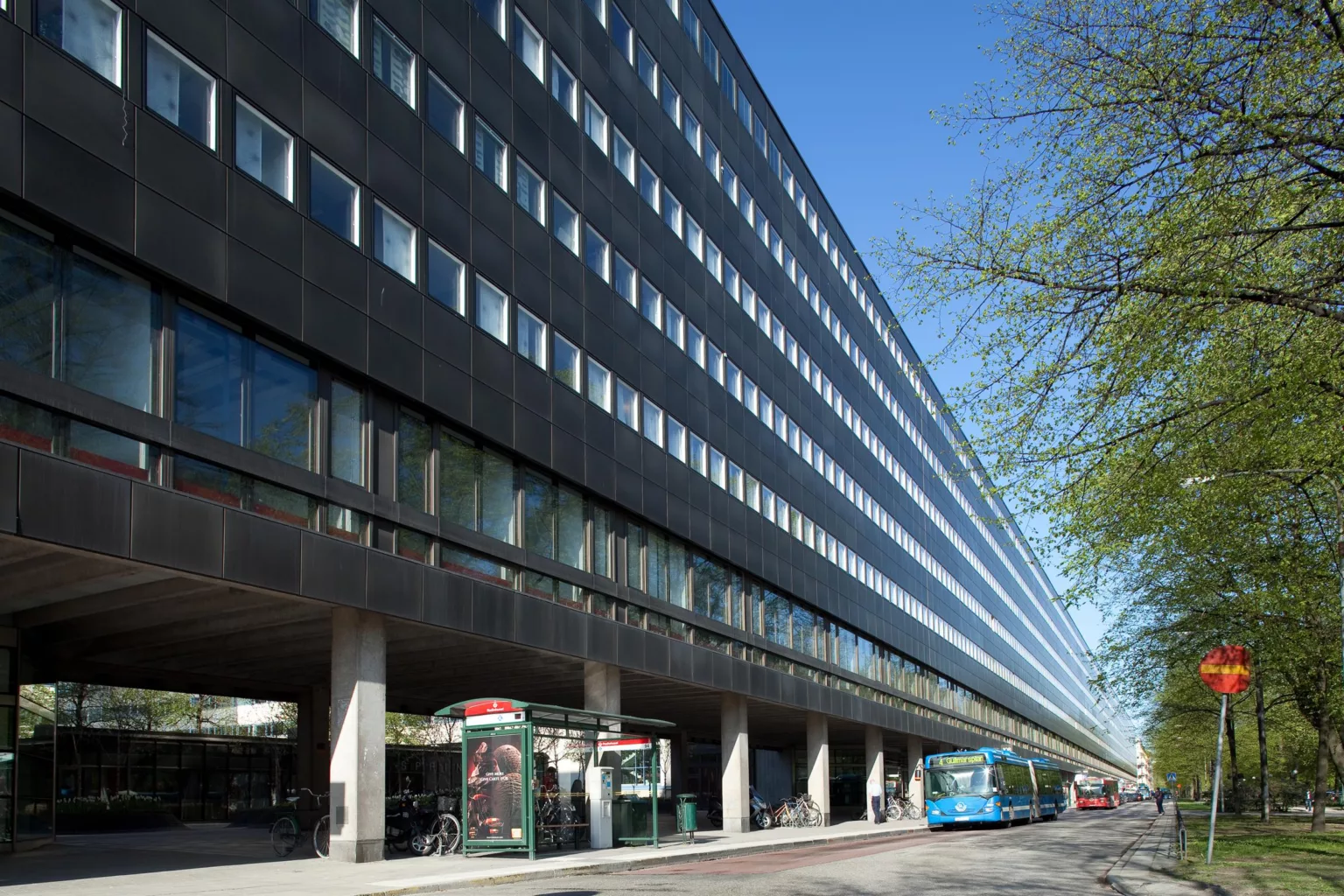 En lång modern byggnad med mörk fasad och glasfönster, med en busshållplats och telefonkiosk framför.
