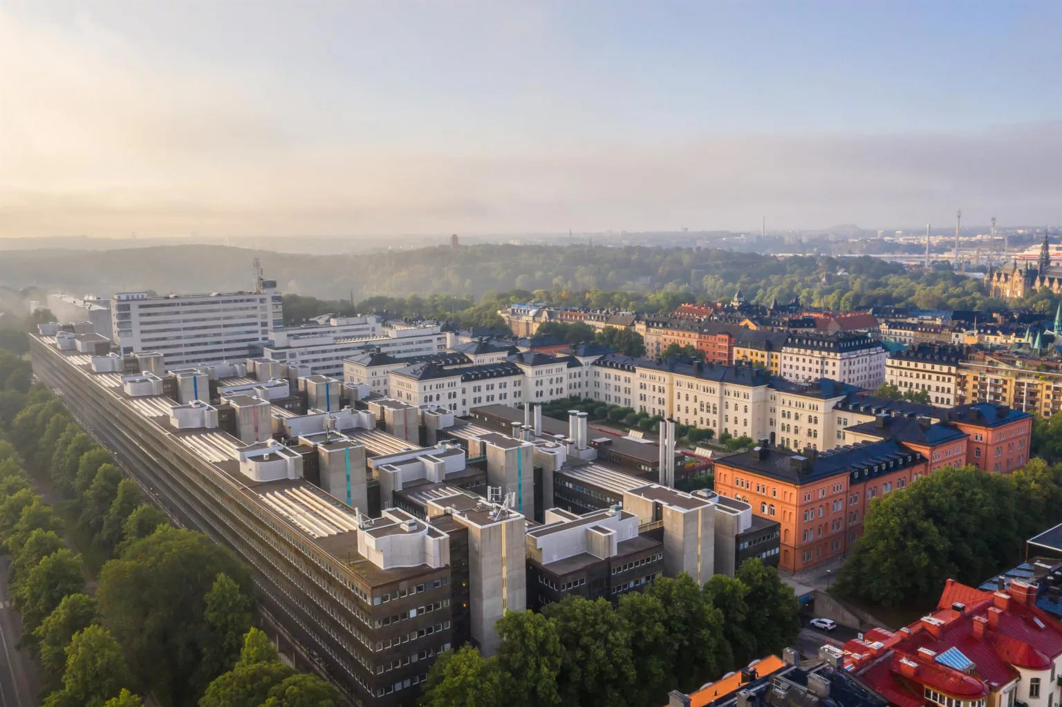Flygbild över moderna byggnader och bostäder i en stadsdel, med grönska och dimmig himmel i bakgrunden.