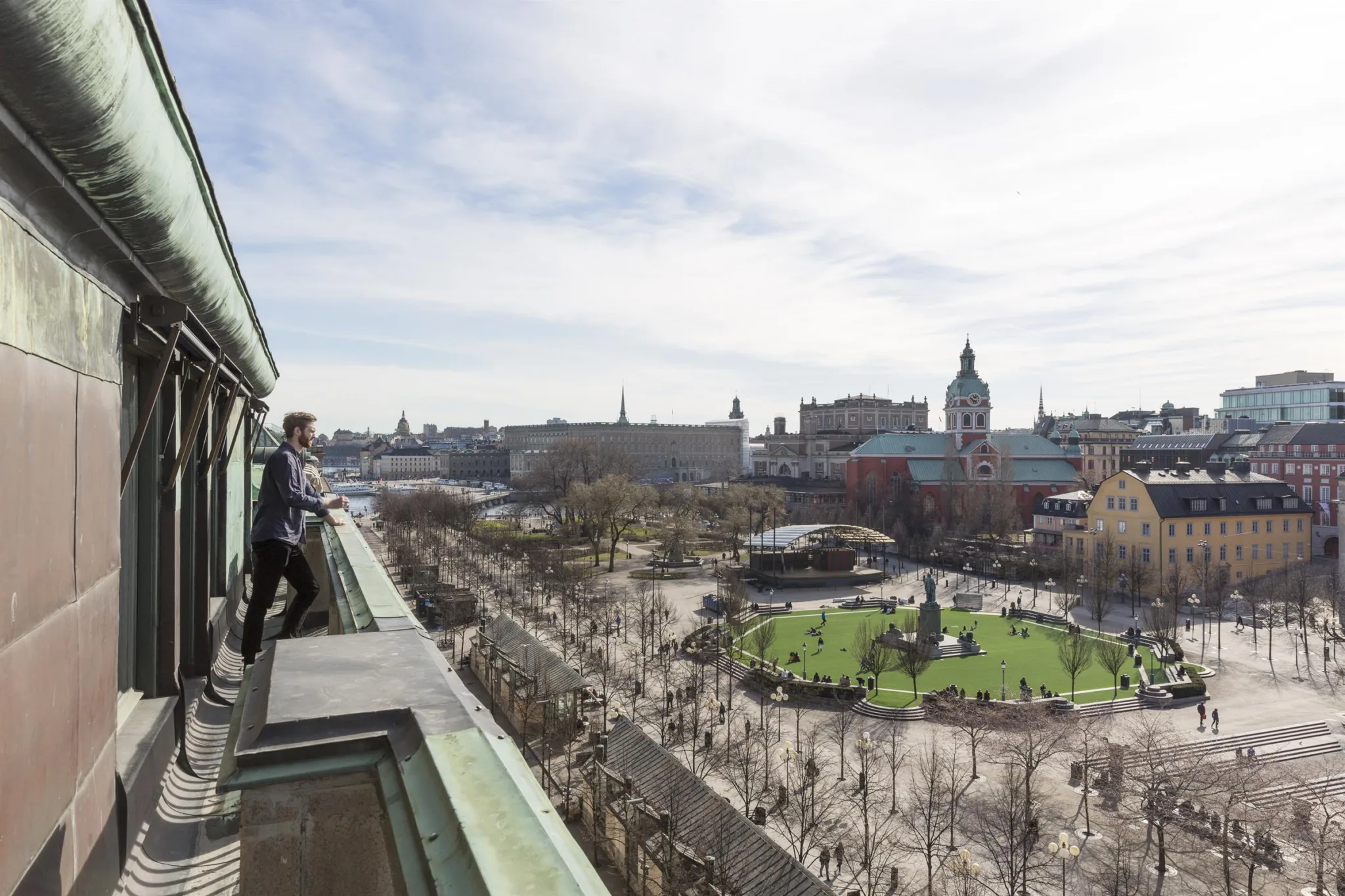 En stadsvy med en park, kyrktorn och personer på en balkong.