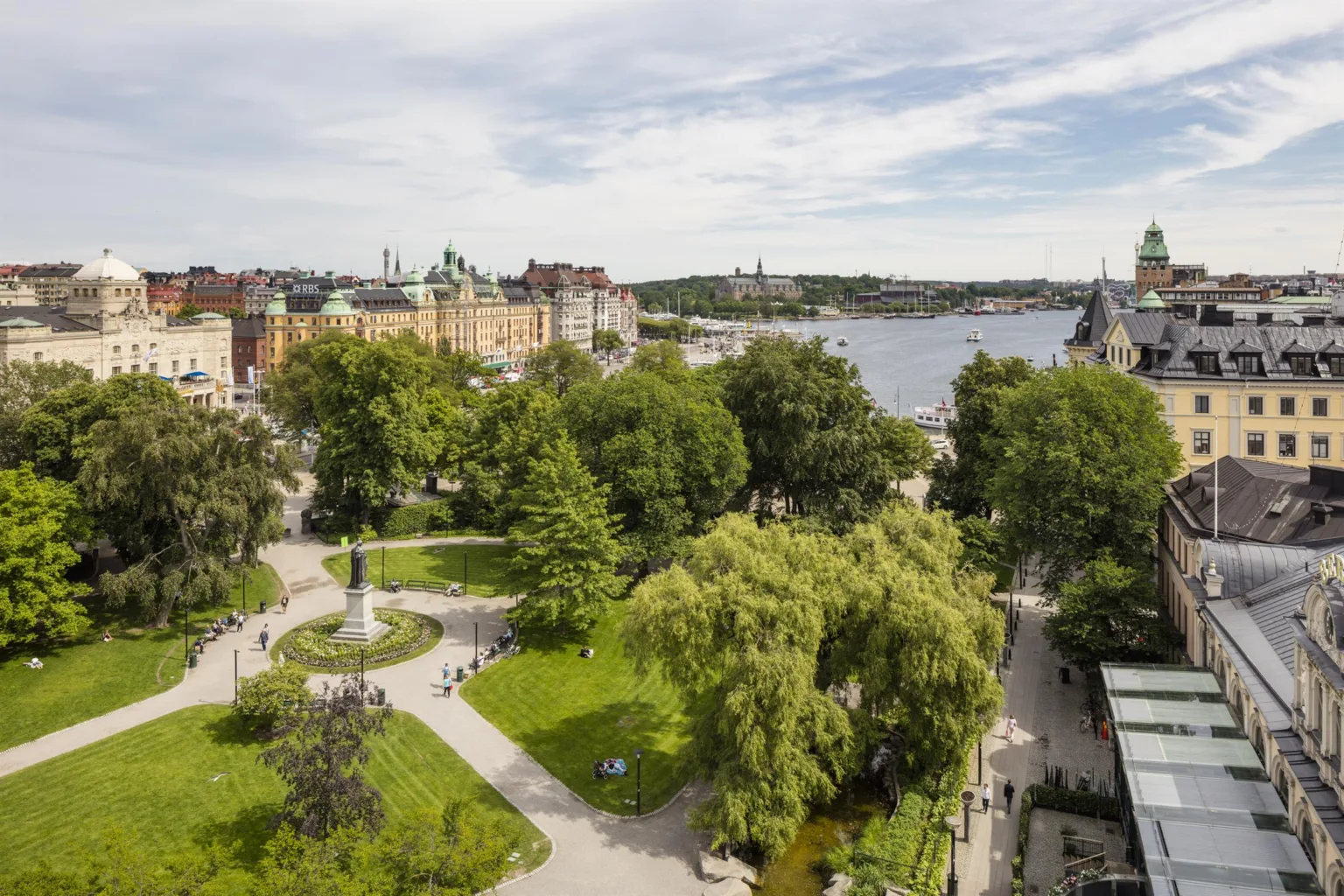 Park med grönska, gångvägar och utsikt över en sjö omgiven av stad och byggnader.