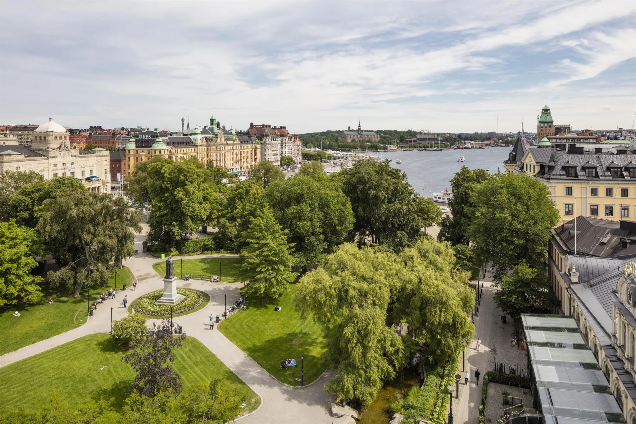 Park med grönska, gångvägar och utsikt över en sjö omgiven av stad och byggnader.