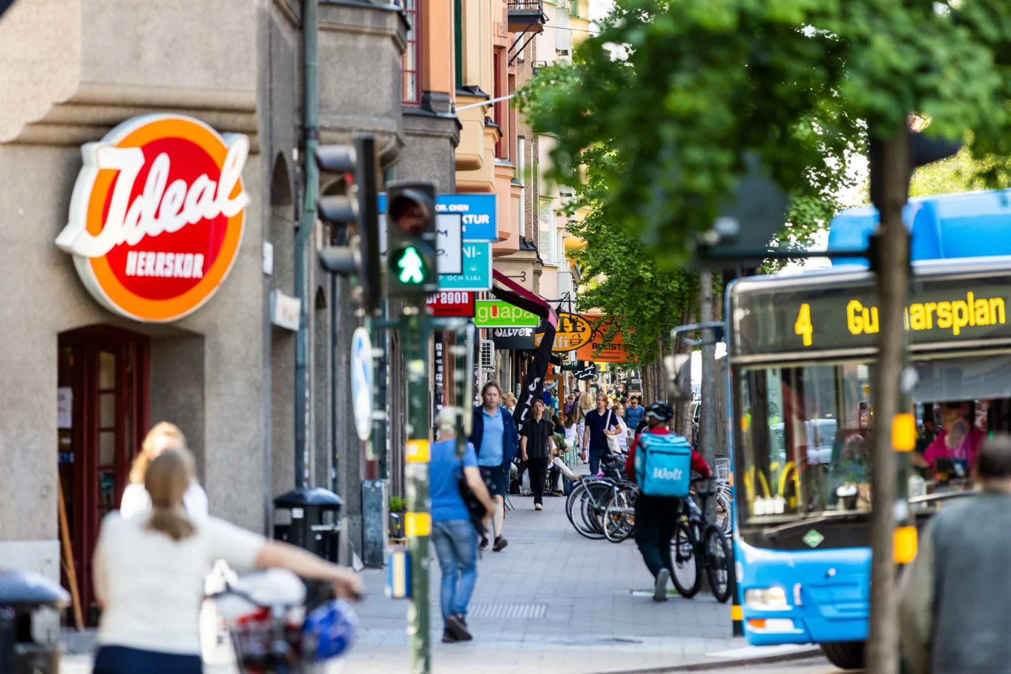 En gata i stadsmiljö med en buss, cyklister och gångtrafikanter samt butiker i bakgrunden.