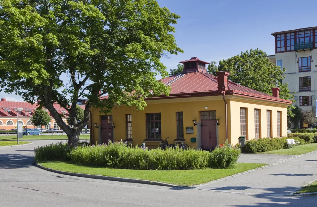 4. En gul byggnad med rödtak och fönster, omgiven av grönska och ett stort träd under en solig himmel.