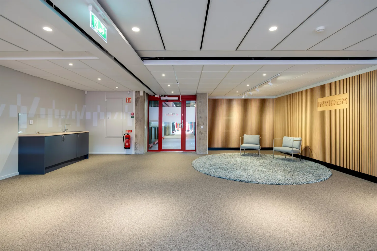 En reception med två fåtöljer på en rund matta, en trävägg, och en glasdörr med röda ramar som leder ut.