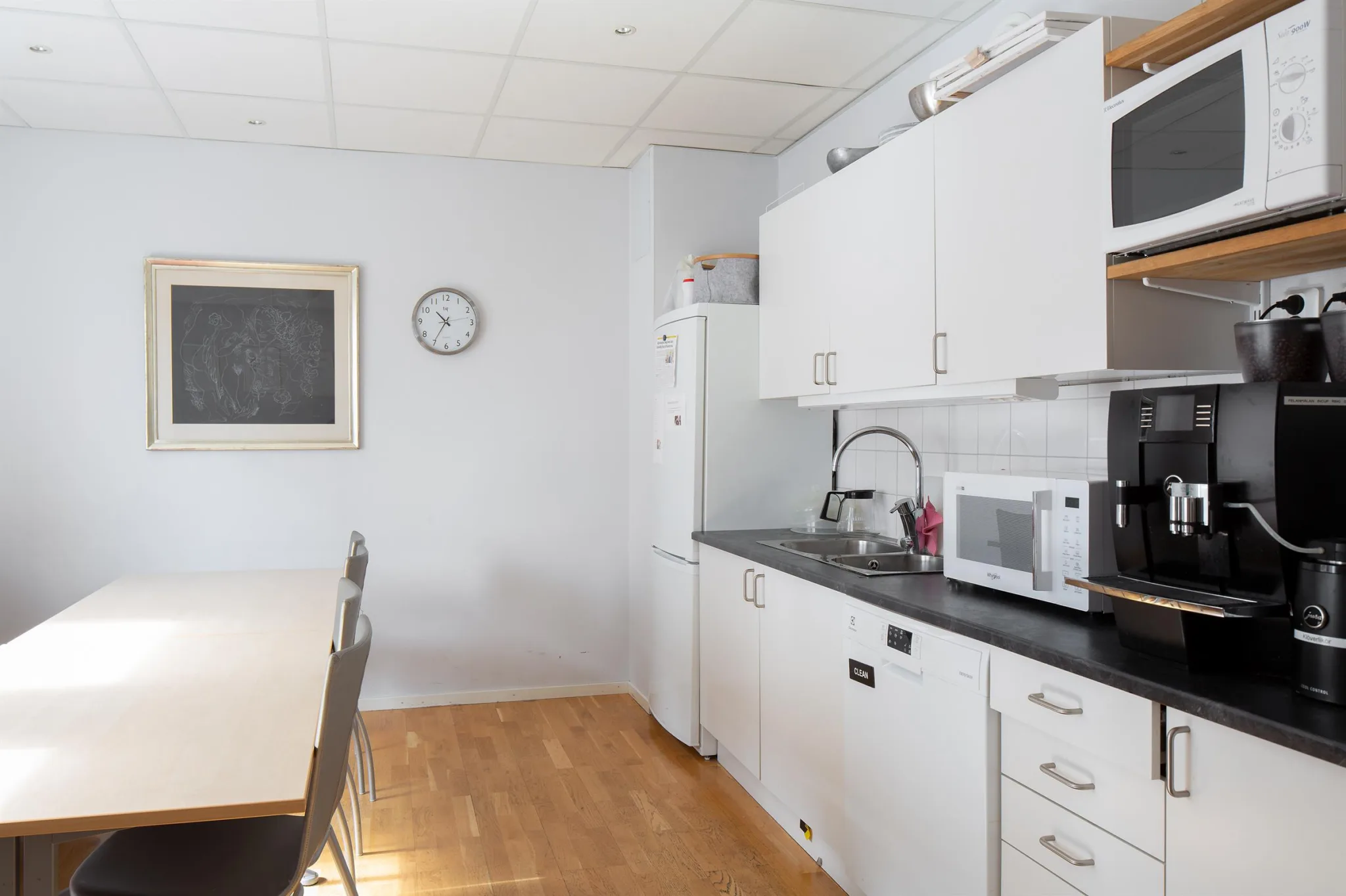 Ett ljust kök med vita skåp, en mikrovågsugn, en klocka på väggen och ett matbord.