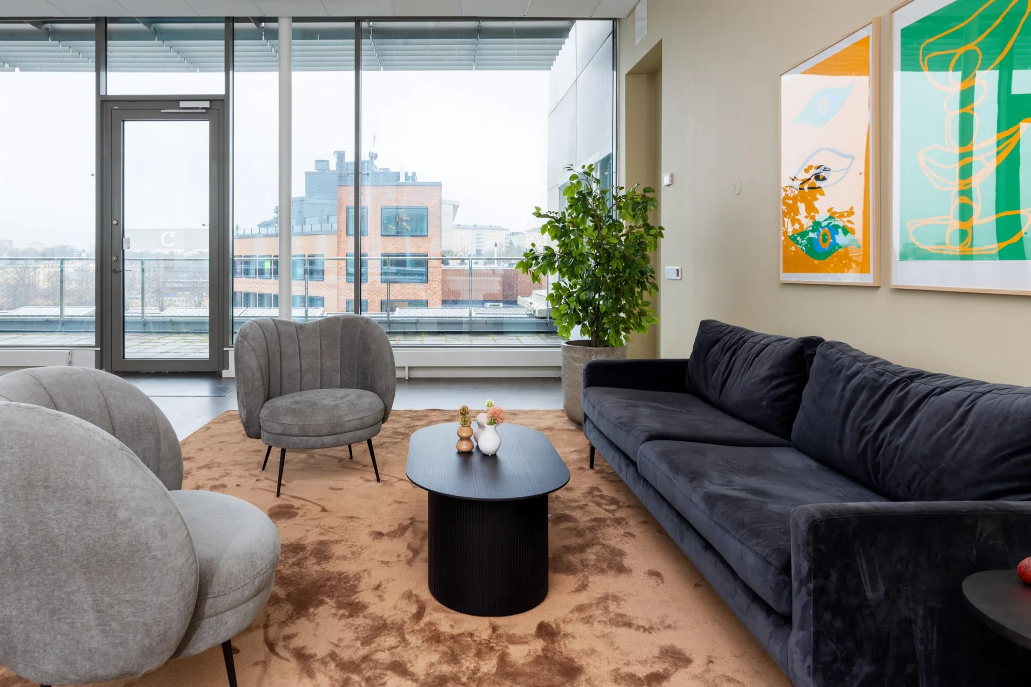 - Ett modernt vardagsrum med grå soffa, två grå fåtöljer, ett svart soffbord, en stor grön växt och en färgglad tavla på väggen. Stora fönster visar utsikt över stadsmiljö.