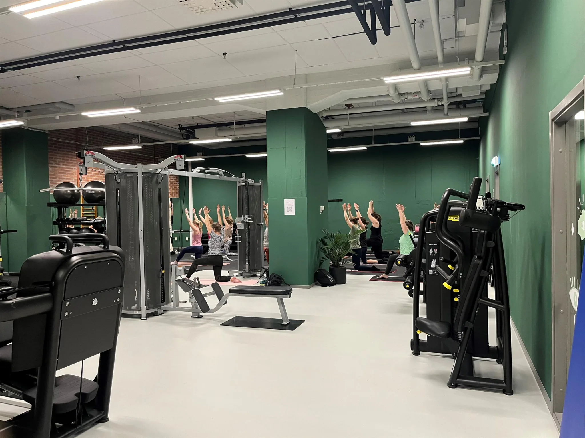 Ett gym med gröna väggar, träningsutrustning och en grupp personer som gör övningar tillsammans.