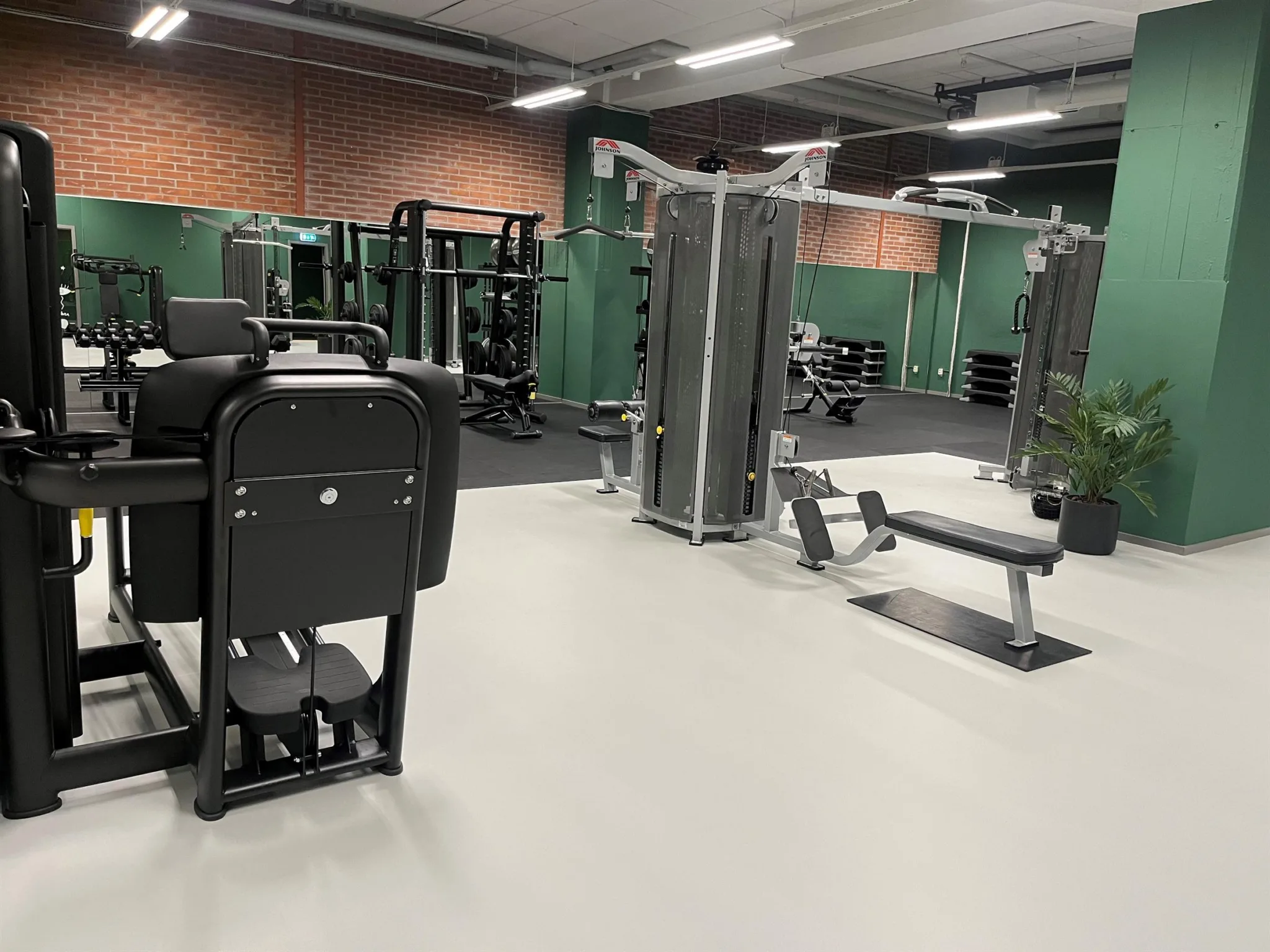 Ett gym med träningsmaskiner, inklusive kabelmaskiner och bänkpress, mot väggar med grön och tegelröd färg.