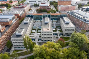 Flygvy över en stor byggnadskomplex med innergårdar och omgivande grönska, flera byggnader och vägar synliga i bakgrunden.