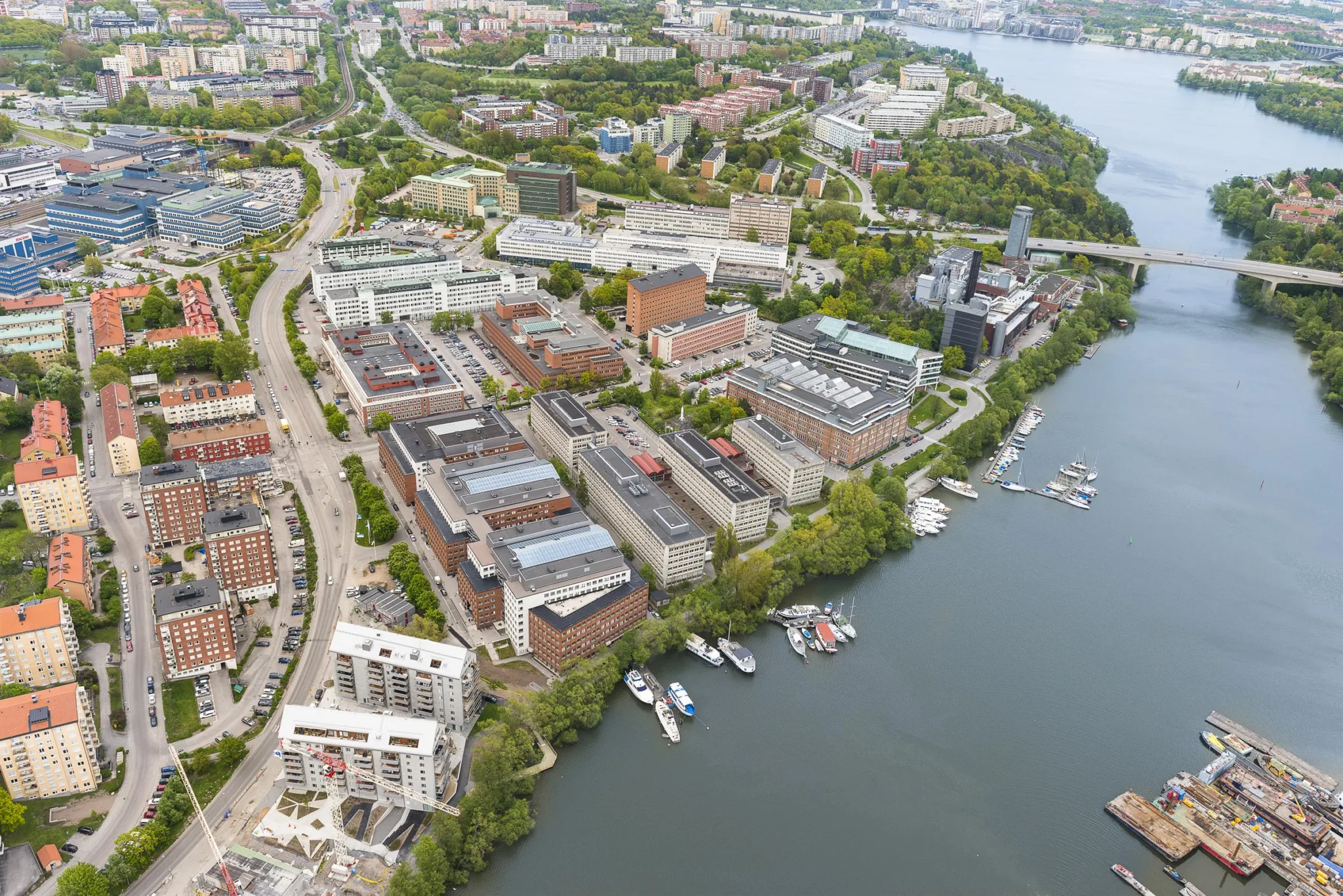 Flygfoto över en stadsdel med byggnader, grönområden och båtar vid en vattensamling.