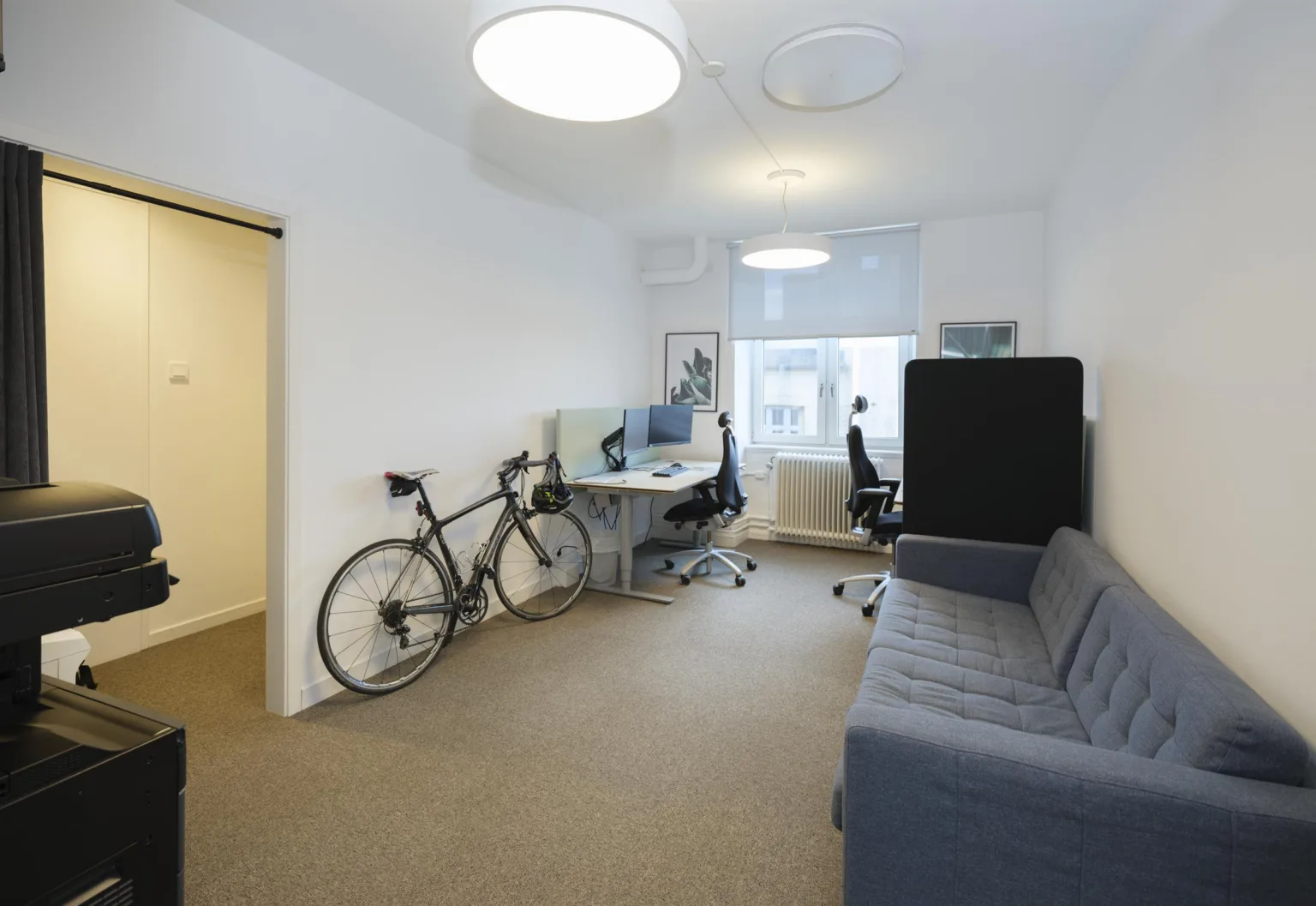 Ett arbetsrum med cykel, skrivbord, stol och en grå soffa, samt stort fönster.