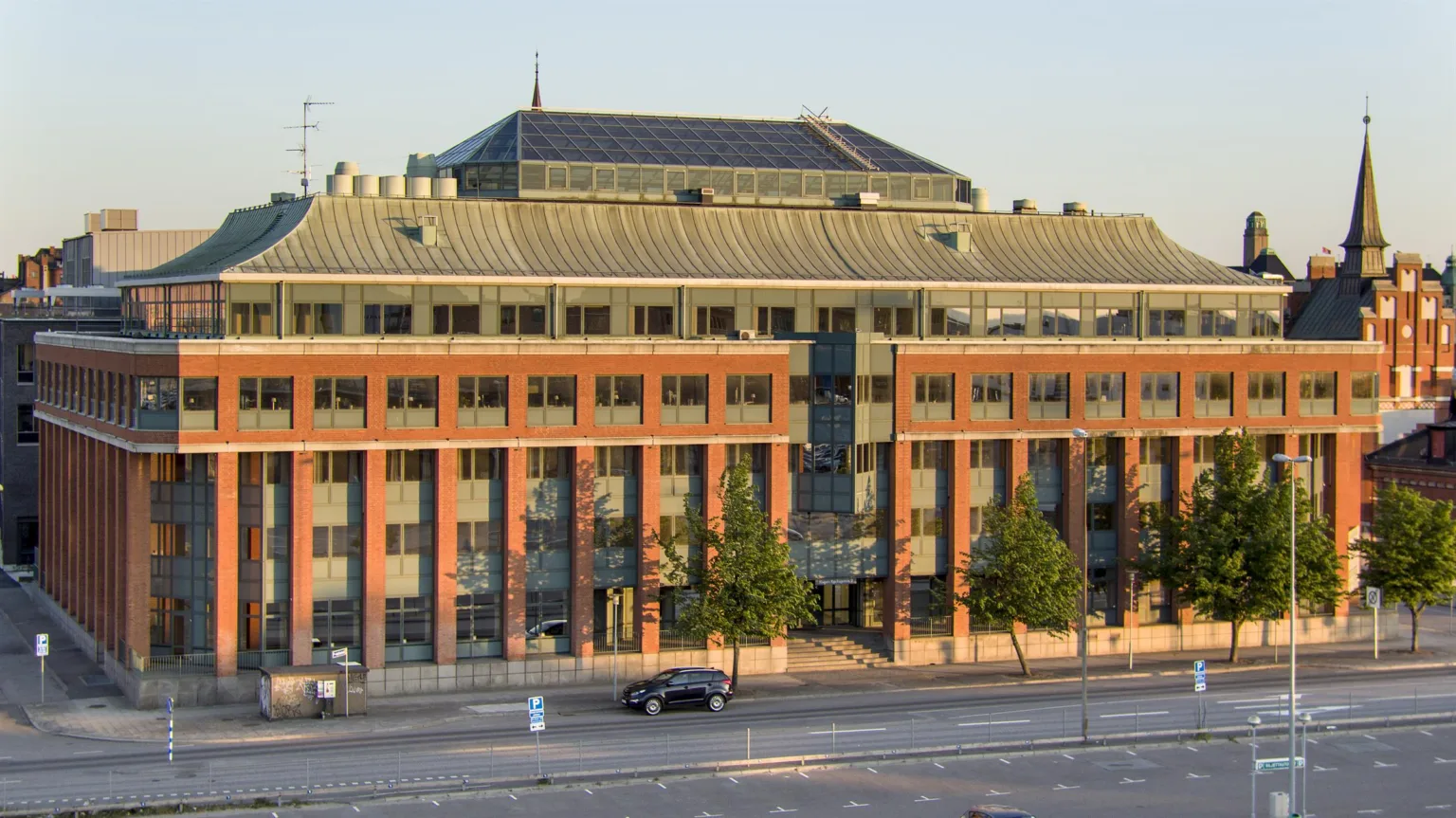 1. En byggnad med rött tegel och stora fönster, omgiven av träd och en gata med en bil.