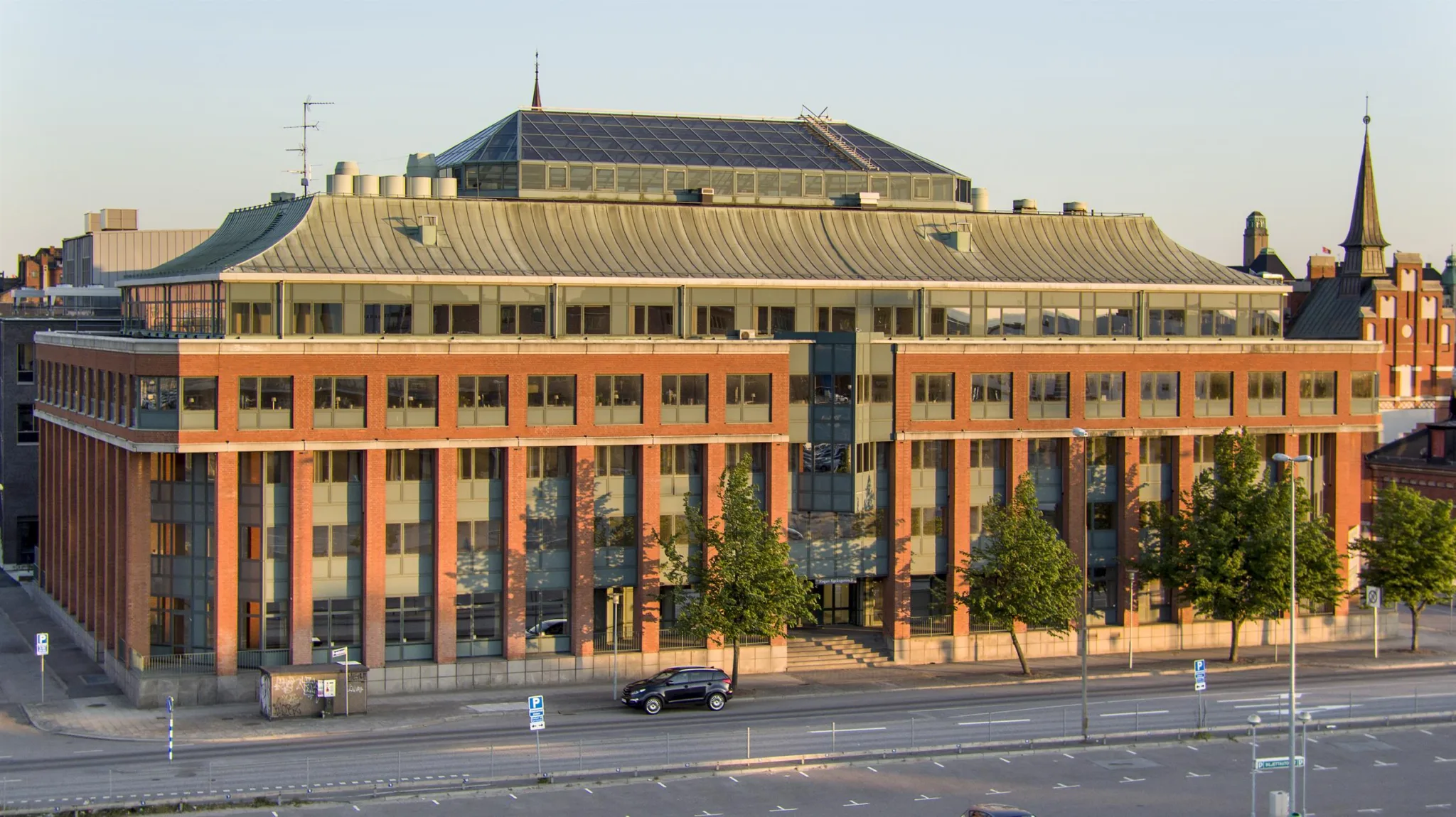 1. En byggnad med rött tegel och stora fönster, omgiven av träd och en gata med en bil.
