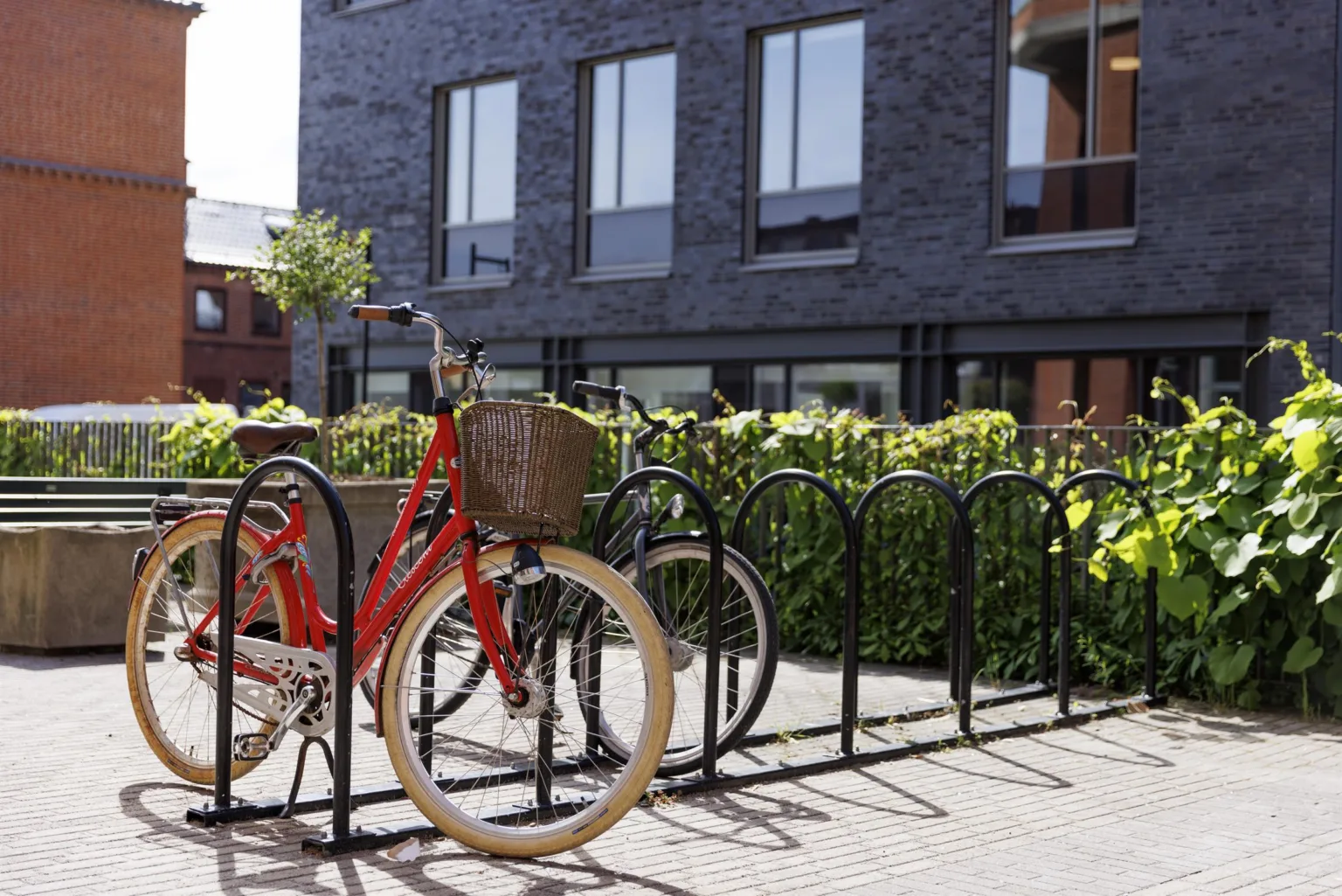 1. En röd cykel med korg står parkerad vid ett cykelställ framför en modern byggnad med stora fönster och gröna buskar.
