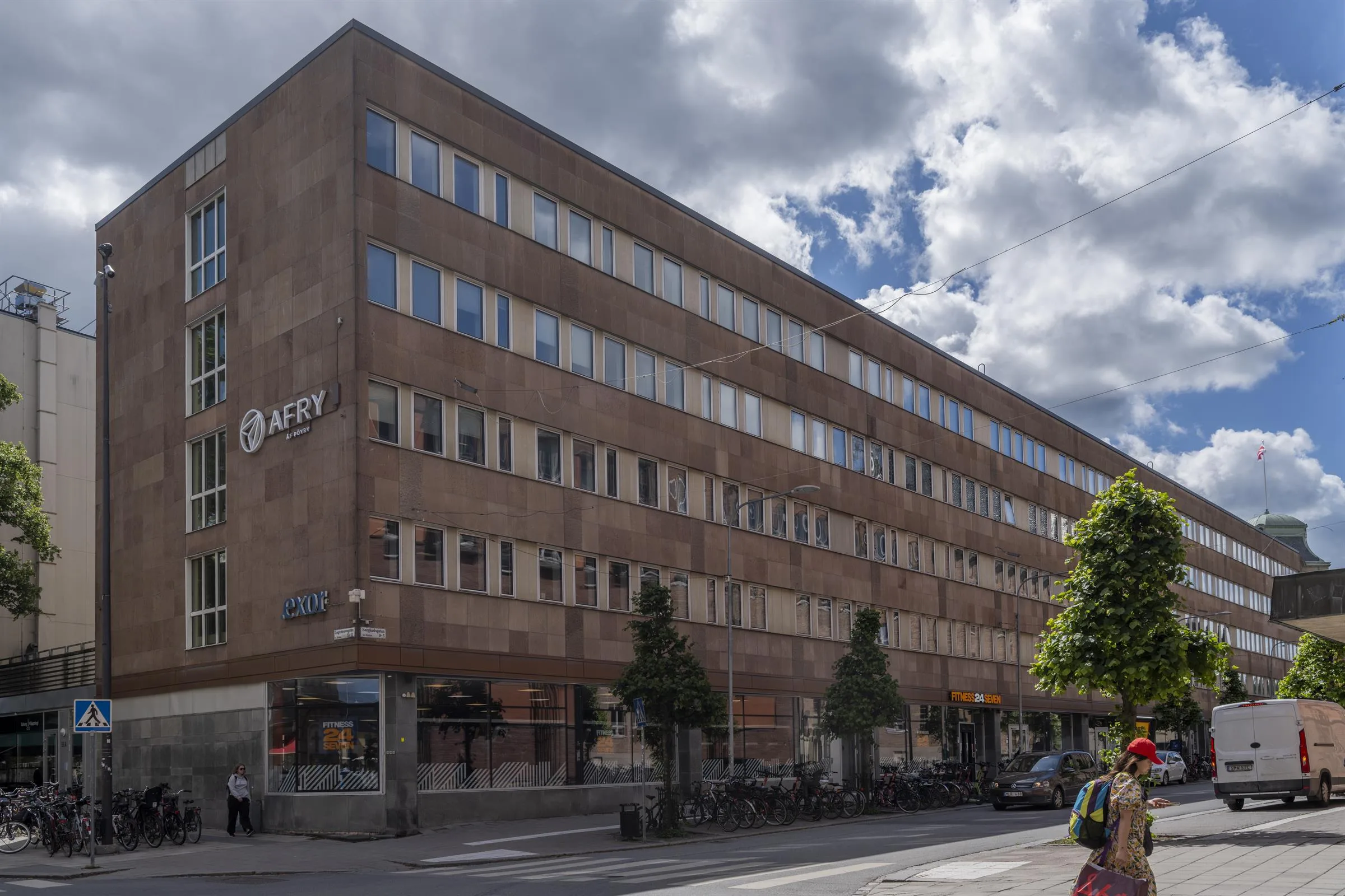 En stor brun tegelbyggnad med flera våningar, fönster och träd längs trottoaren. Skyltar är synliga på byggnadens fasad.