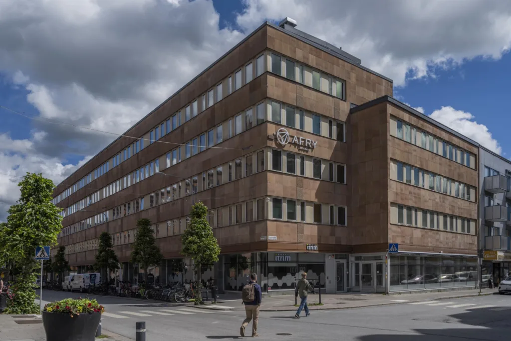 En tegelbyggnad med flera våningar, fönster och en skylt som visar namnet på byggnaden. Människor går på trottoaren nedanför.