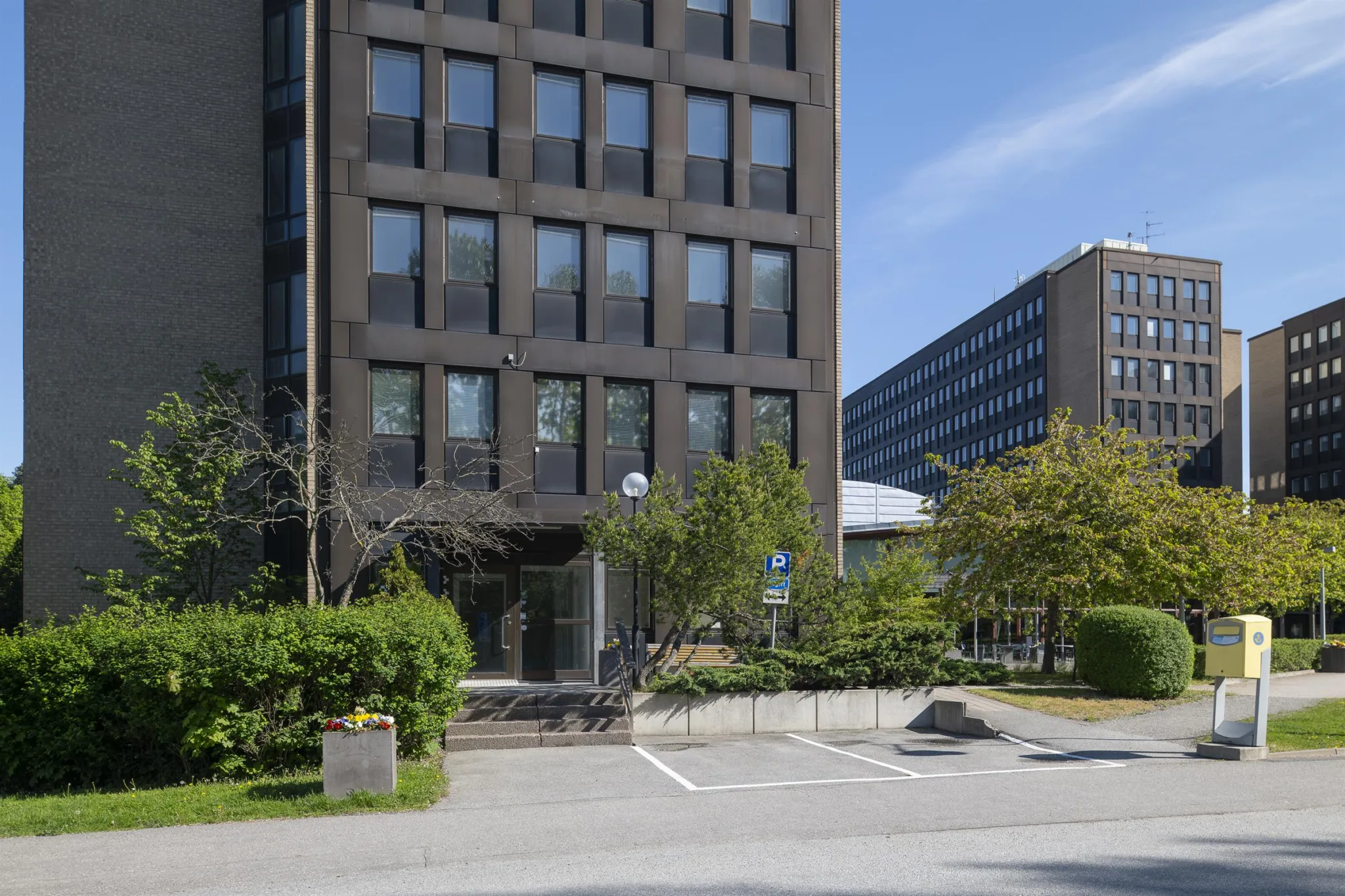 Byggnad med mörk fasad och stora fönster, omgiven av grönska och en parkeringsplats.